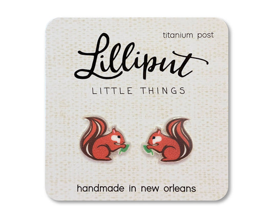 The Red Squirrel Earrings feature two adorable red squirrel shapes holding green leaves, displayed on a card labeled Lilliput Little Things. Handmade in New Orleans with hypoallergenic titanium posts, they are perfect for sensitive ears.