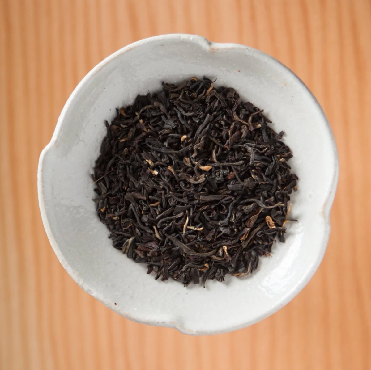 A white, scalloped ceramic bowl filled with Coastal Breakfast (ORG) – Loose Leaf Tin tea, viewed from directly above on a light wood surface.