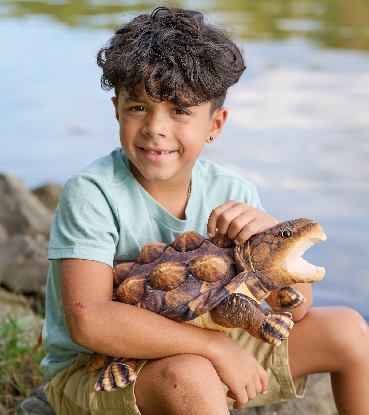 A young boy sits by the water with a big smile, holding the Wild Onez Snapping Turtle 15″ (38cm) plush toy. Surrounded by rocks and lush greenery, this scene offers charming inspiration for swamp-themed nursery decor.