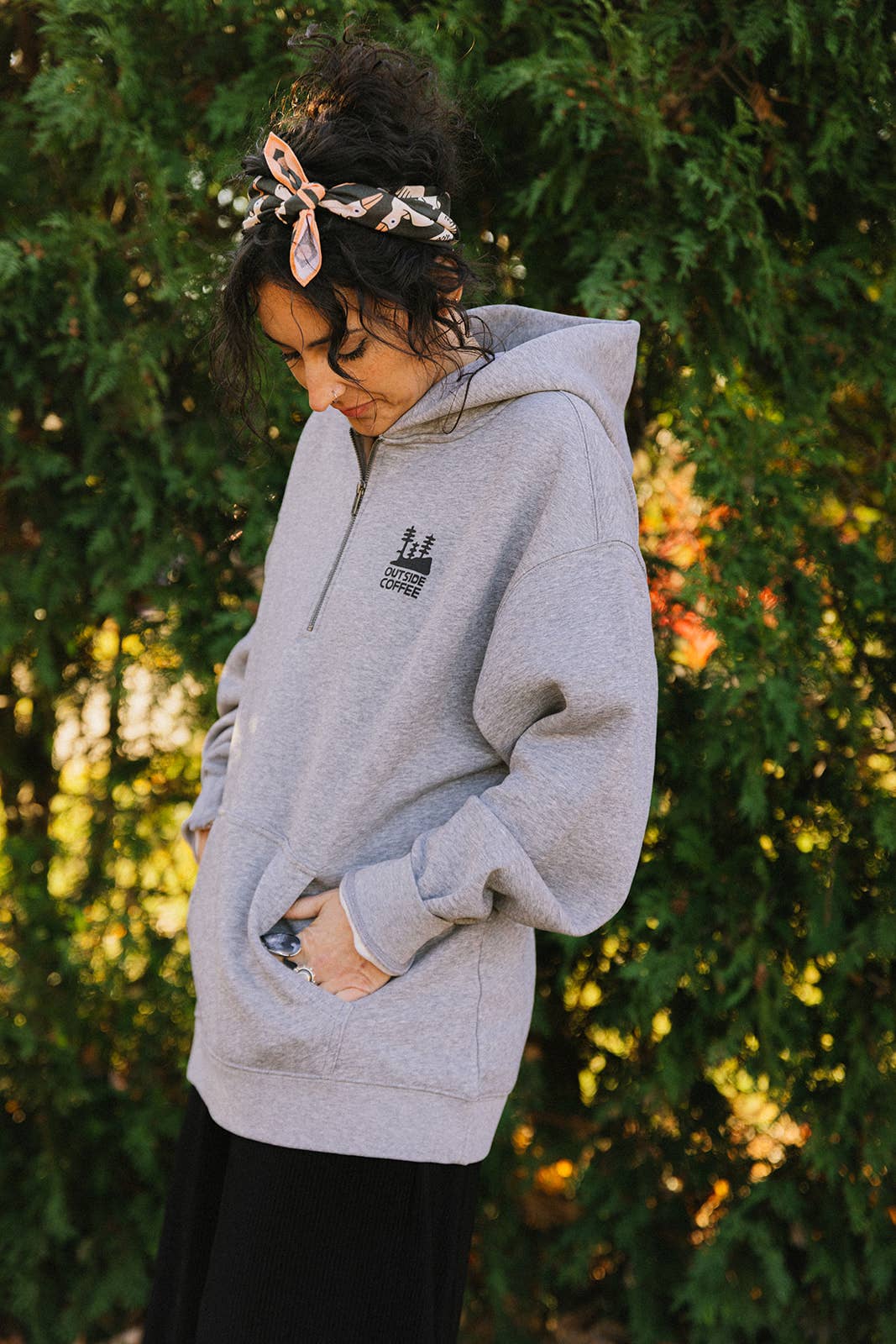 A woman with dark hair tied up in a Migration Bandana, featuring a vibrant sublimated print, stands outdoors in front of green bushes wearing a gray hoodie with her hands in the pockets and looking down.
