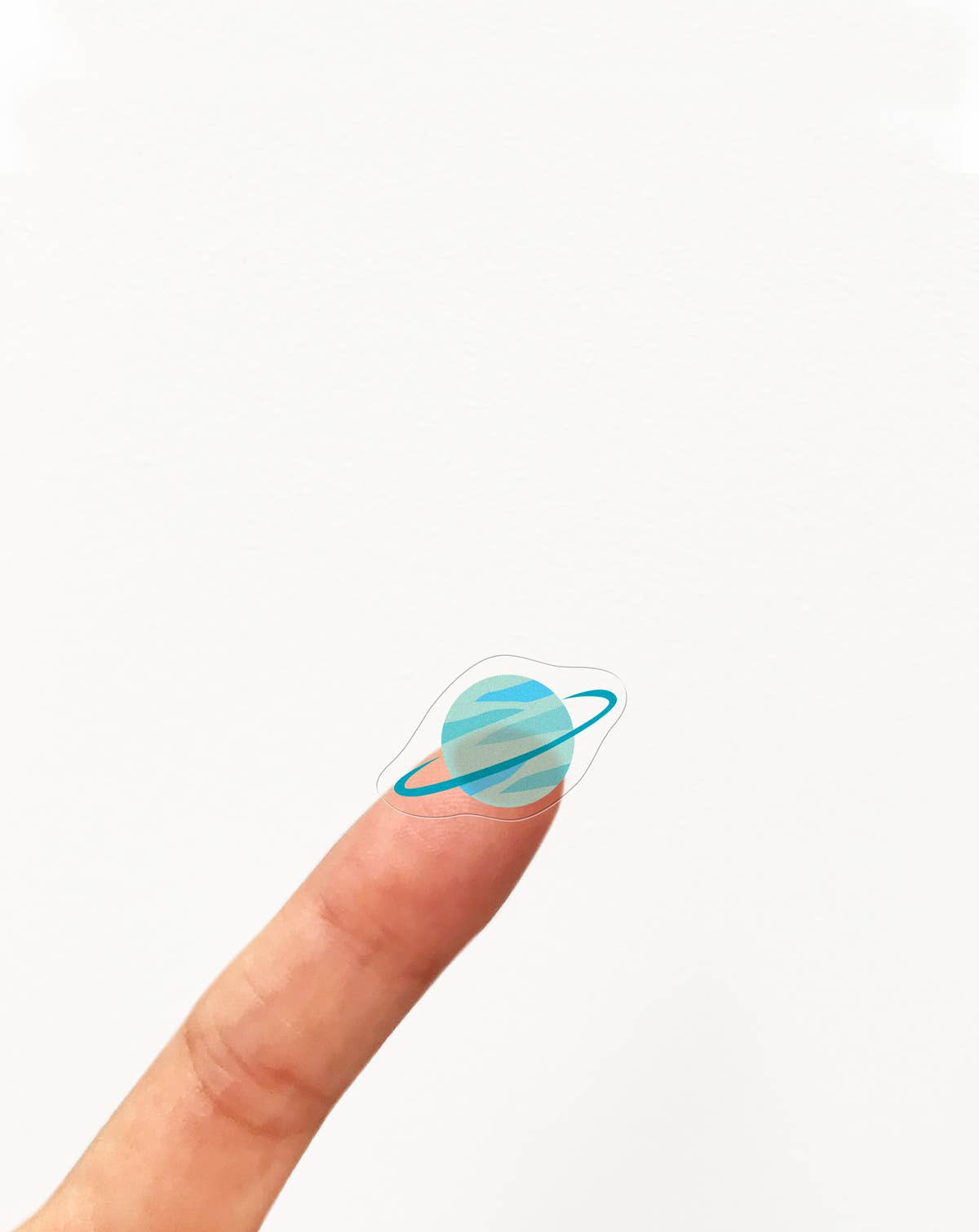 A hand holds a Planet Clear Sticker with a blue and green ringed planet, resembling Saturn, making it a stylish addition to any space-themed collection. The sticker is displayed against a plain white background.
