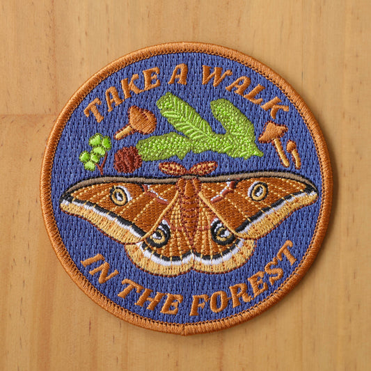 The Take a Walk in the Forest Iron-on Patch is circular and embroidered with a moth, leaves, and mushrooms. It has orange text on blue, an orange border, and is shown on a wooden surface.