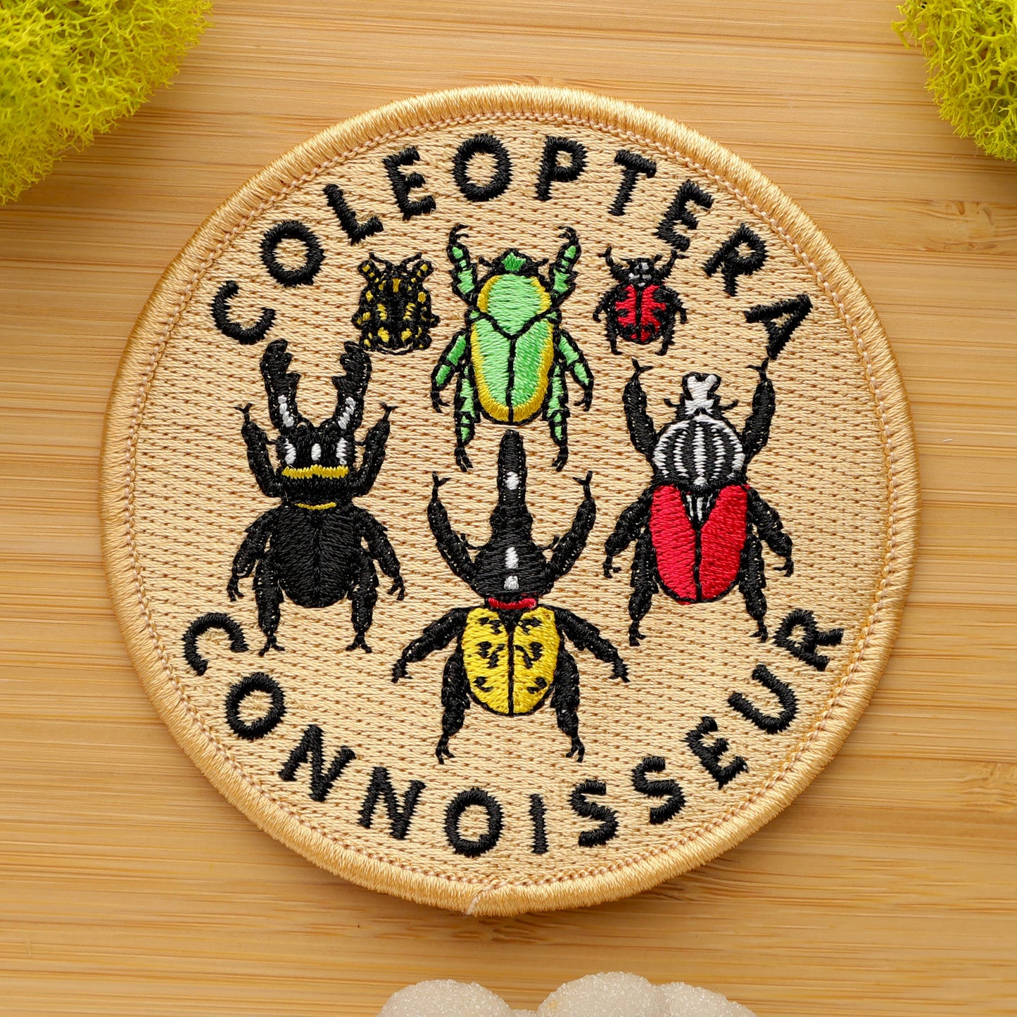 The Coleoptera Connoisseur Iron-on Patch is a round, intricately embroidered patch featuring six colorful beetles and the phrase “Coleoptera Connoisseur” on a light tan background. It’s shown on wood with yellow and white sponges nearby.