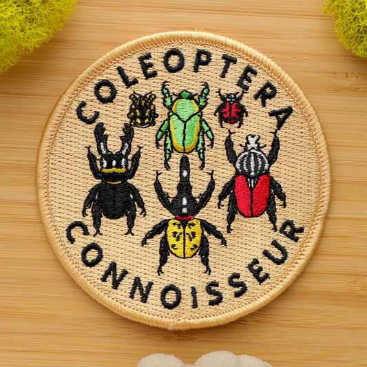 The Coleoptera Connoisseur Iron-on Patch is a round, intricately embroidered patch featuring six colorful beetles and the phrase “Coleoptera Connoisseur” on a light tan background. It’s shown on wood with yellow and white sponges nearby.