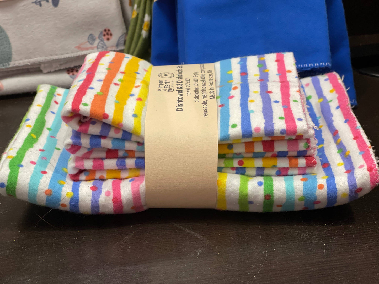 The Dishtowel & 3 Dishcloth Set–Compostable features colorful cotton flannel towels with stripes and dots, wrapped in a beige paper band, displayed on a dark surface with blue and white fabrics in the background.