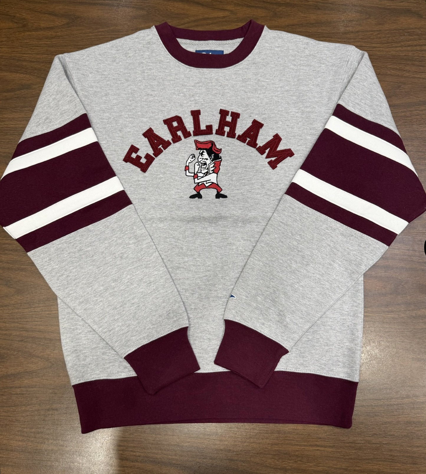 A Fighting Quaker Retro Crew in gray with maroon cuffs and sleeve stripes, "EARLHAM" in maroon above a mascot graphic on the chest, is shown laid flat on a wooden surface.