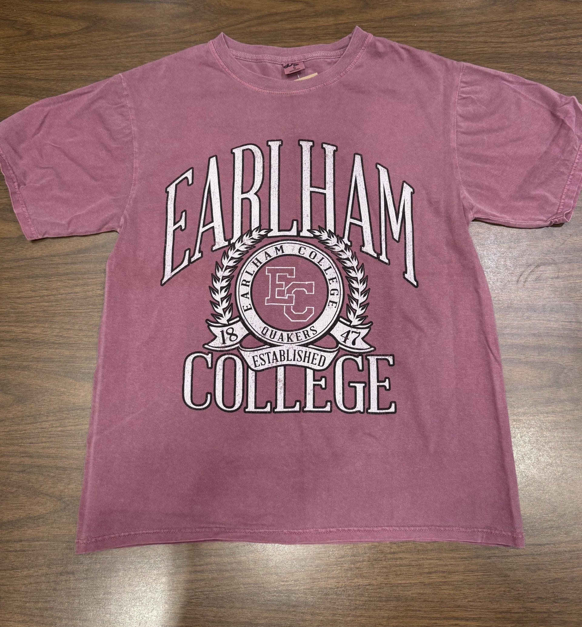 A Vintage Wreath Tee in faded maroon, featuring Earlham College in bold white letters, a central EC Quakers crest, and "Established 1847" below, displayed flat on a wooden surface.