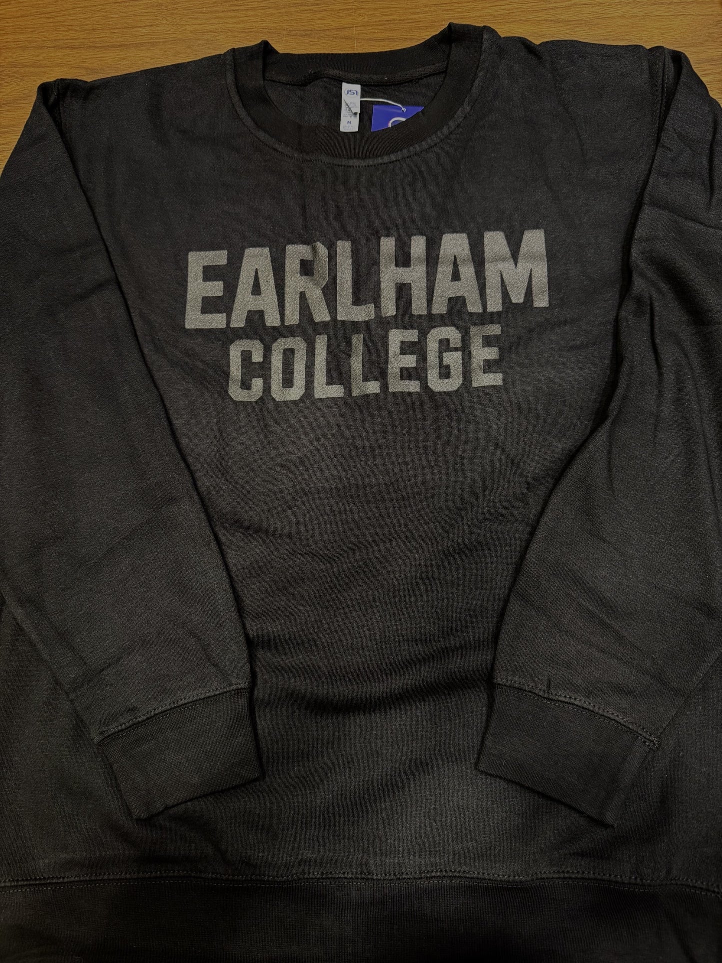 A Black Tonal Puff Crew sweatshirt featuring "EARLHAM COLLEGE" in large, gray block letters across the chest, displayed flat on a wooden surface.
