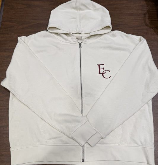 A French Terry Zip-Up Oversized Hoodie in cream, featuring a hood and maroon "EC" lettering on the left chest, is laid flat on a wooden surface.