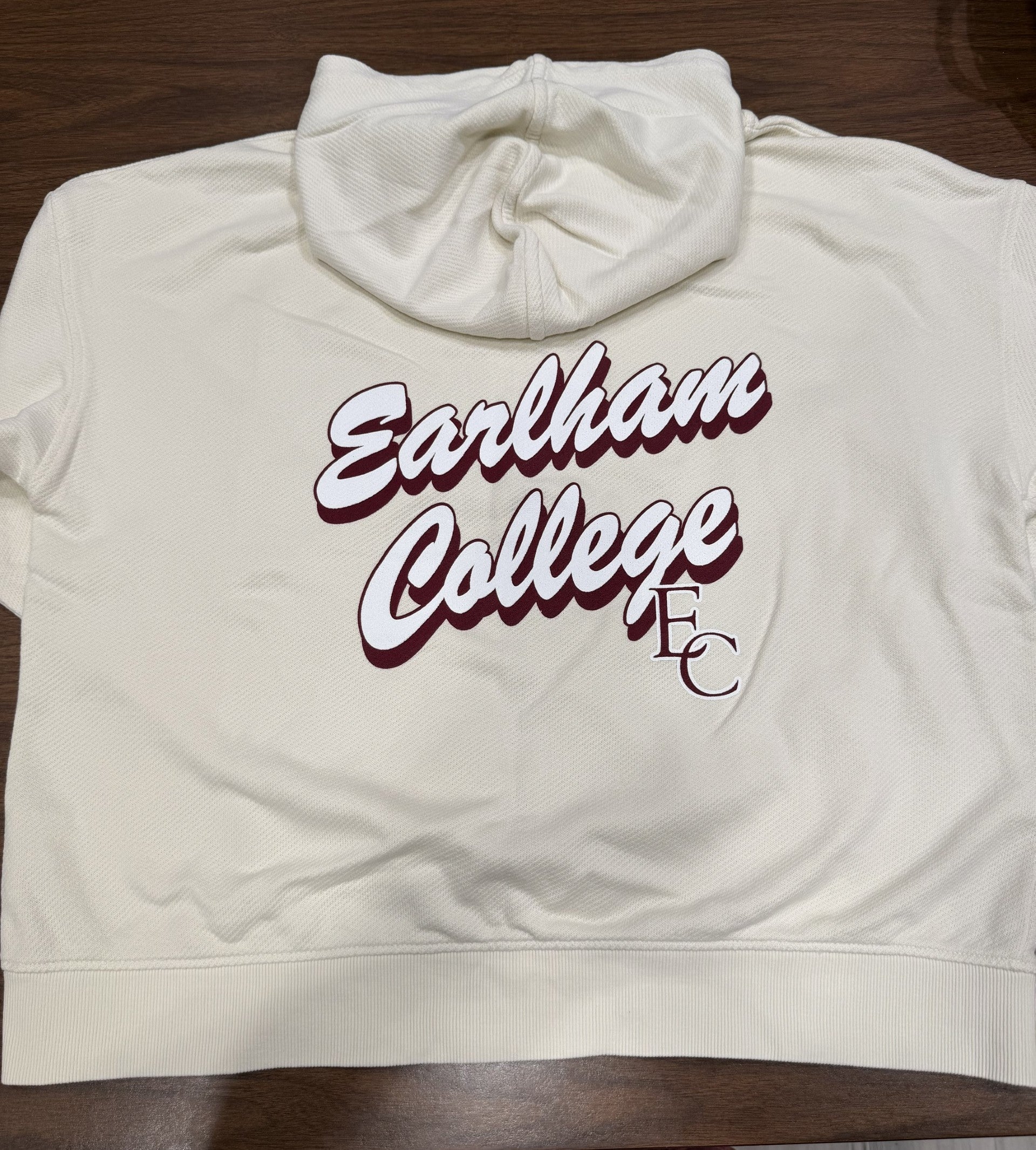 A French Terry Zip-Up Oversized Hoodie in cream, featuring maroon "Earlham College" text on the front and "EC" on the back, displayed flat on a wooden surface.