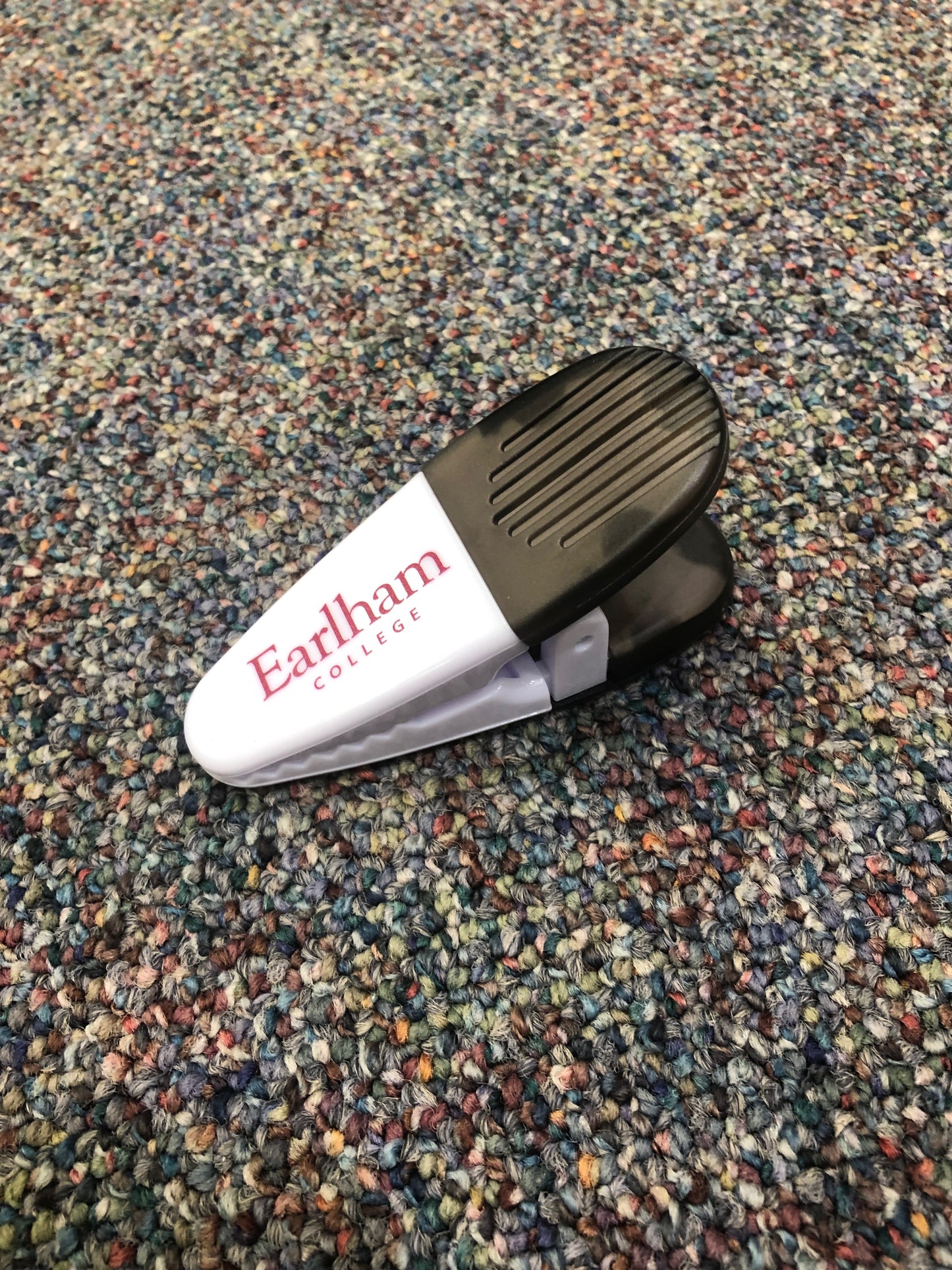 A Magnetic Chip Clip in white and black, featuring "Earlham College" printed in red, rests on a multicolored speckled carpet.