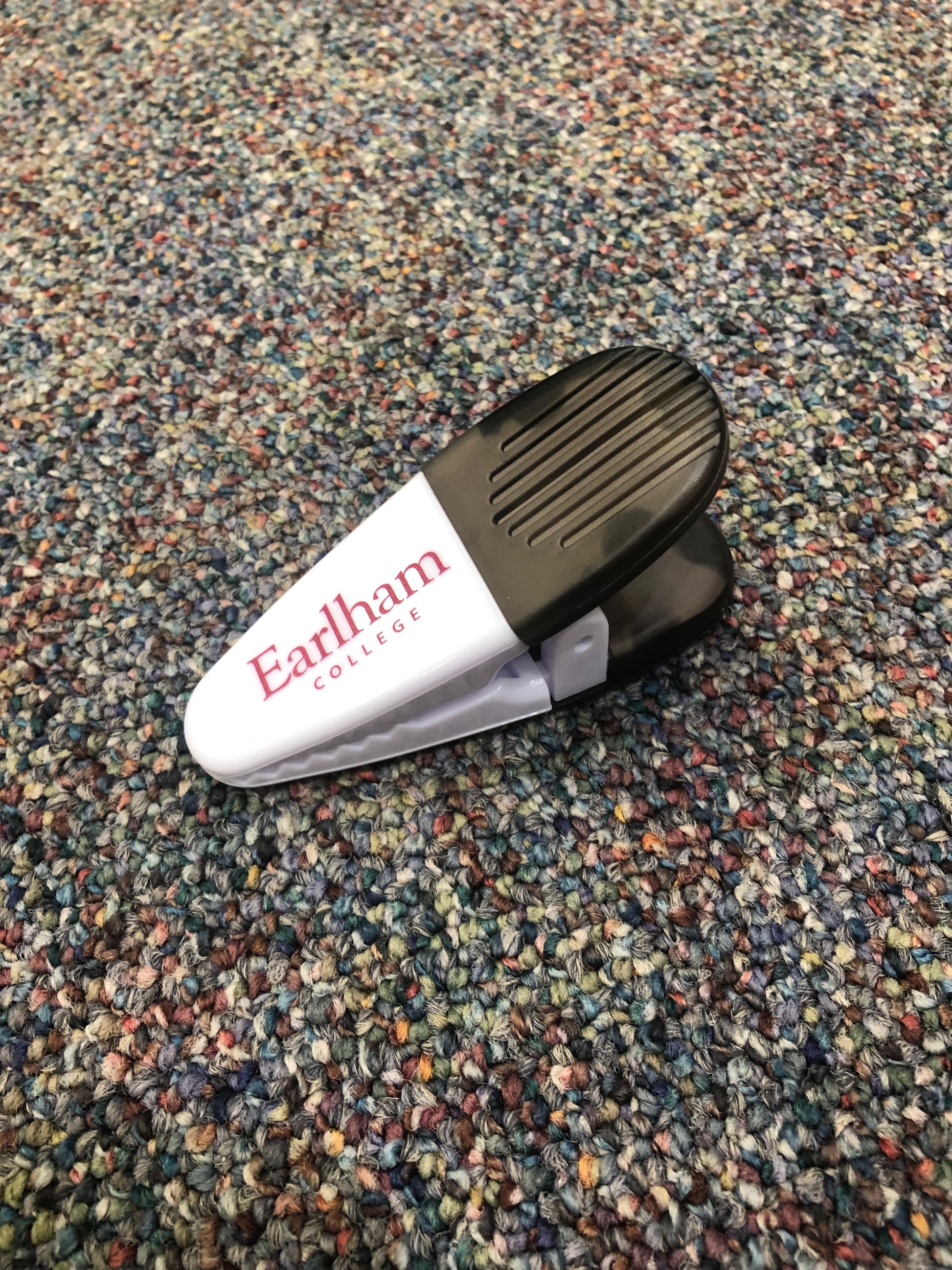 A Magnetic Chip Clip in white and black, featuring "Earlham College" printed in red, rests on a multicolored speckled carpet.