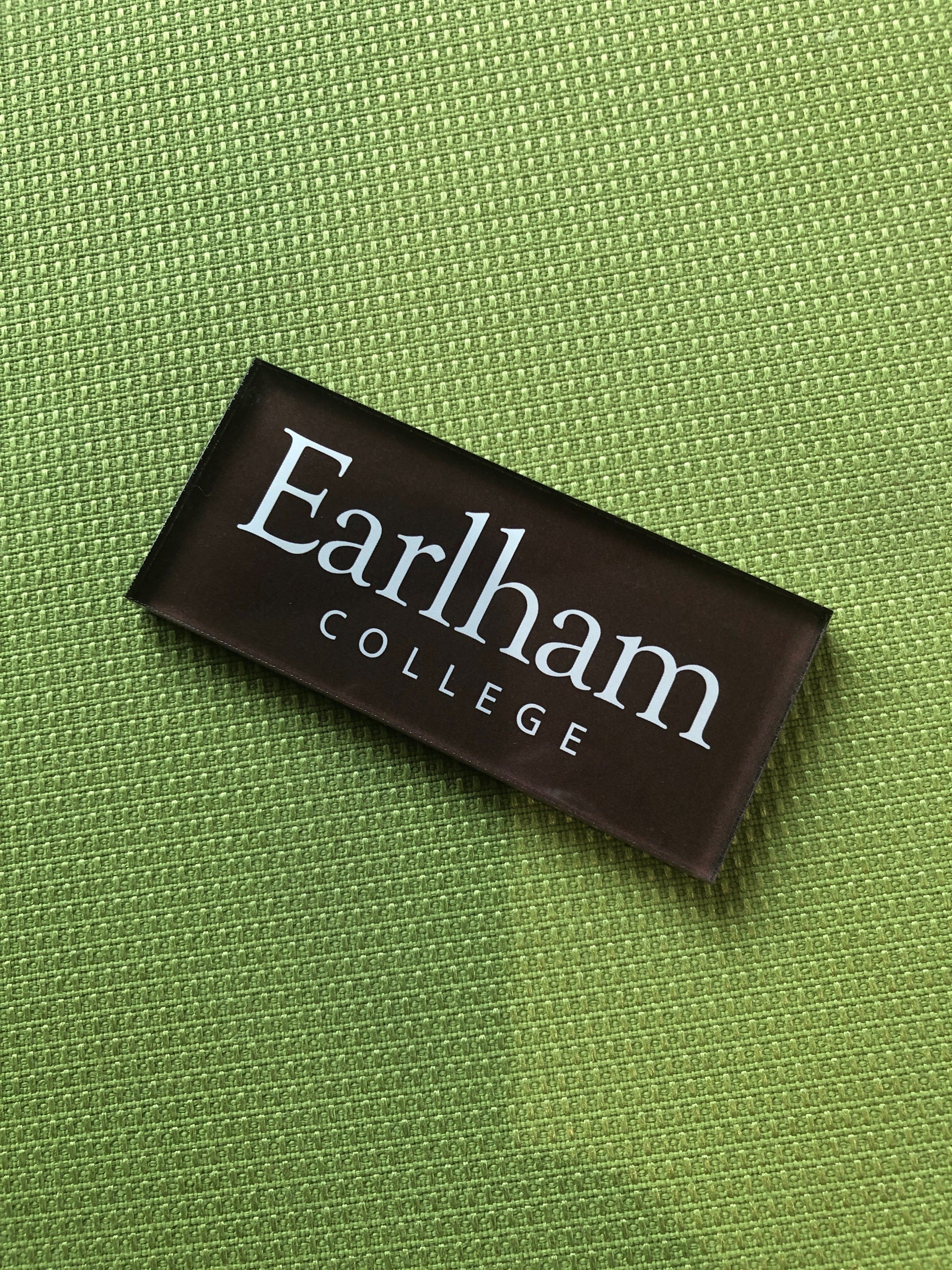 A rectangular black Earlham Acrylic Refrigerator Magnet with "Earlham College" in white text, placed on a textured green surface.