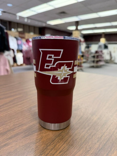 The Maroon Travel Tumbler 20oz, featuring a white and gold EC logo with a starburst design and silver base, sits on a wooden table with clothing displays and shelves in the background.