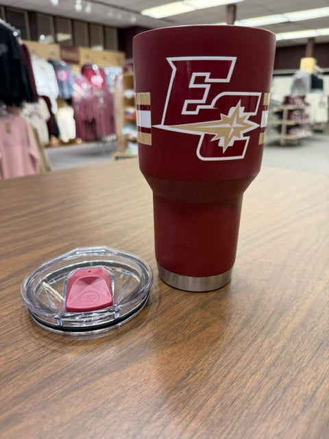 The Maroon Travel Tumbler 30oz, featuring a white and gold EC logo, rests on a wooden table with its clear lid and red slider beside it. Clothing racks and shelves are seen in the background.