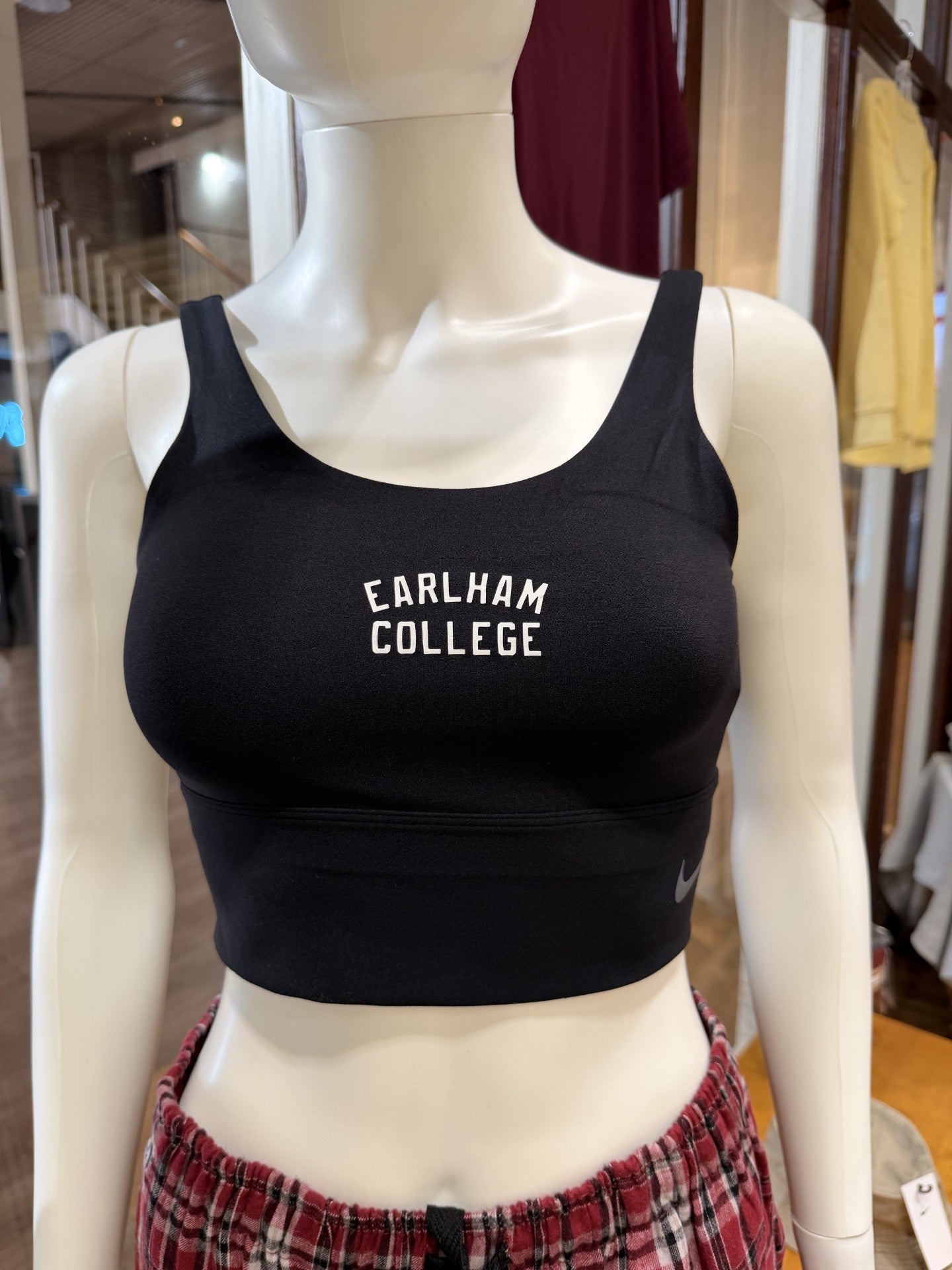 A mannequin wears a black Nike Sports Bra with EARLHAM COLLEGE printed on the front and plaid pants. The background shows a store interior with clothing and wooden accents.
