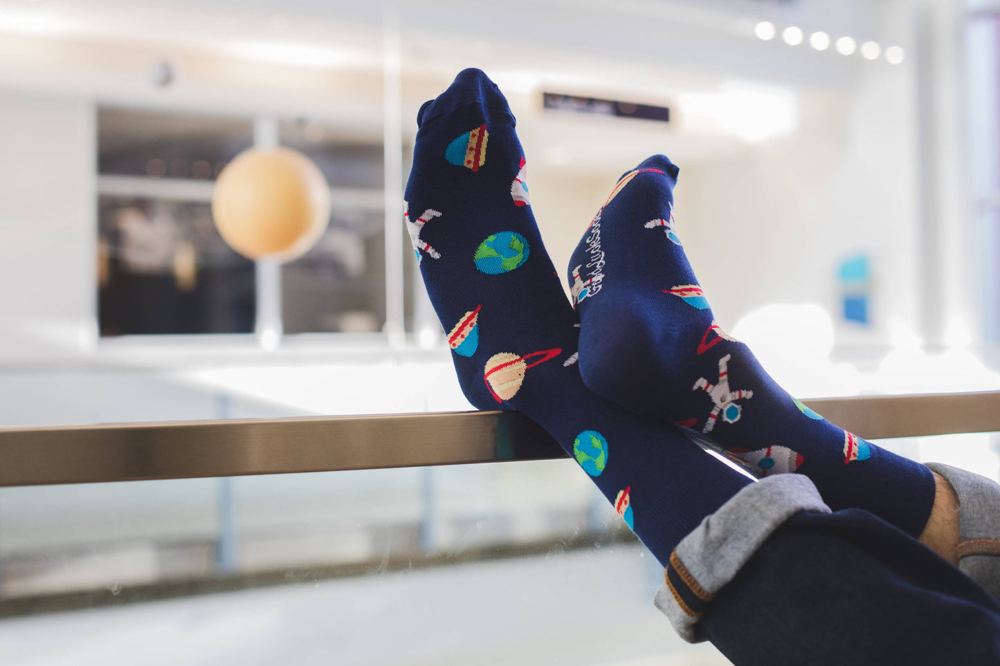 A person wearing Men's Space Socks with colorful planet and rocket designs rests their feet on a glass railing in a bright, modern indoor space.