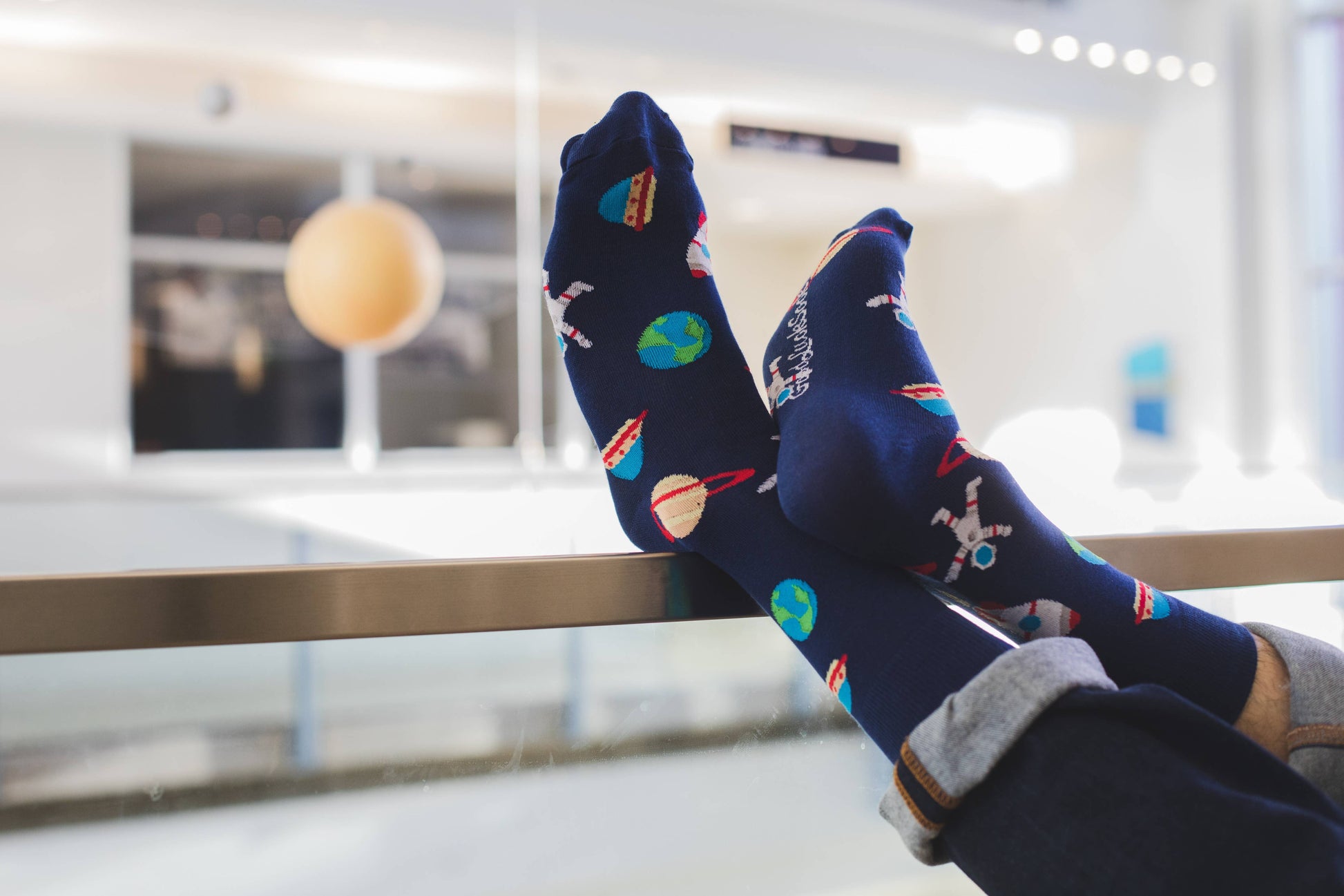 A person wearing Men's Space Socks with colorful planet and rocket designs rests their feet on a glass railing in a bright, modern indoor space.