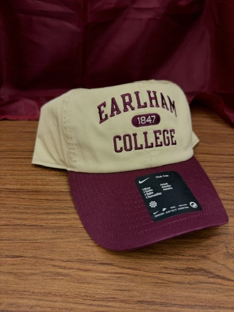 A beige and maroon Nike Club Colorblock Cap with 1847 displayed on the front rests on a wooden surface. A black Nike tag is attached to the brim, and a maroon cloth is draped in the background.