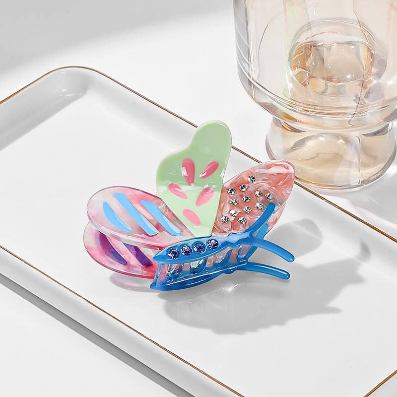 Side Butterfly Hair Claw clip featuring multicolored wings and embedded rhinestones on a white tray.