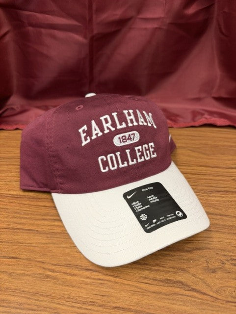 A Nike Club Colorblock Cap in maroon and white, featuring EARLHAM 1847 COLLEGE embroidered on the front, rests on a wooden surface with a maroon fabric backdrop.