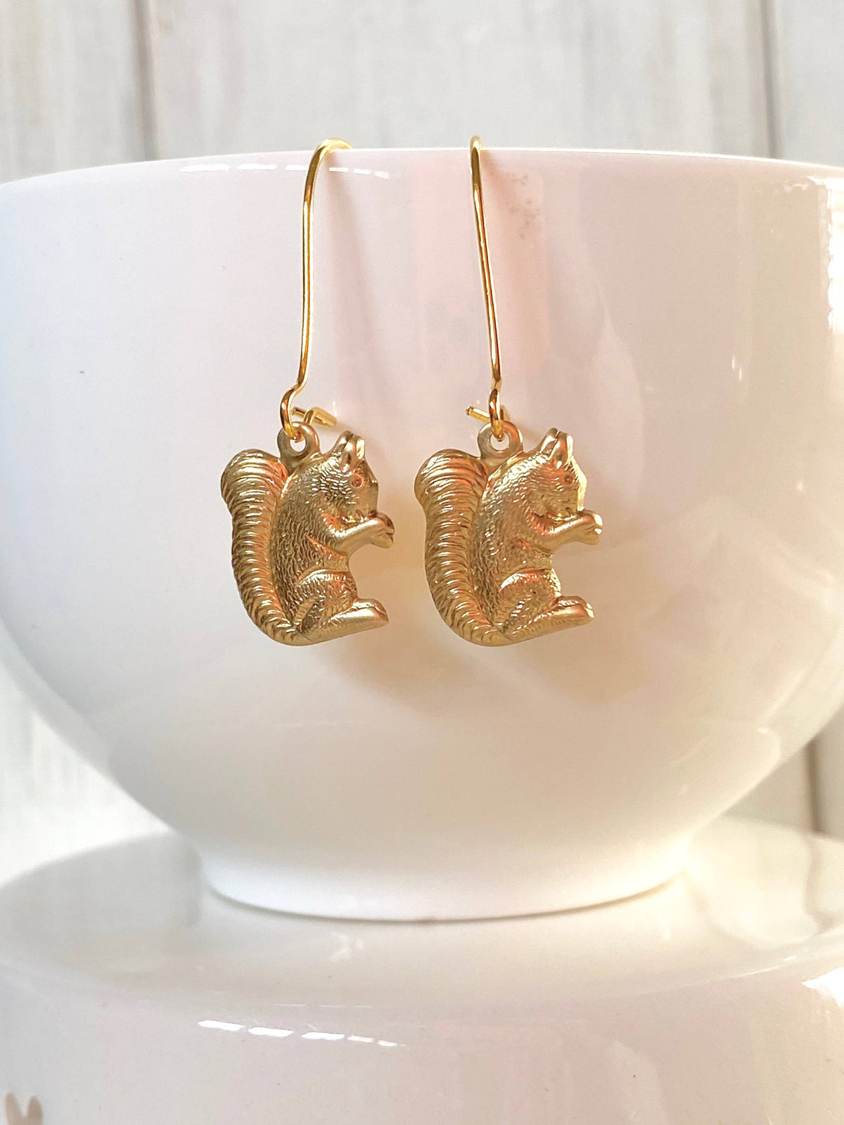 Woodland Forest Squirrel Earrings with gold squirrel-shaped charms and hooks, displayed on the rim of a white ceramic bowl against a soft, light background—delightful woodland-inspired jewelry perfect for nature lovers.
