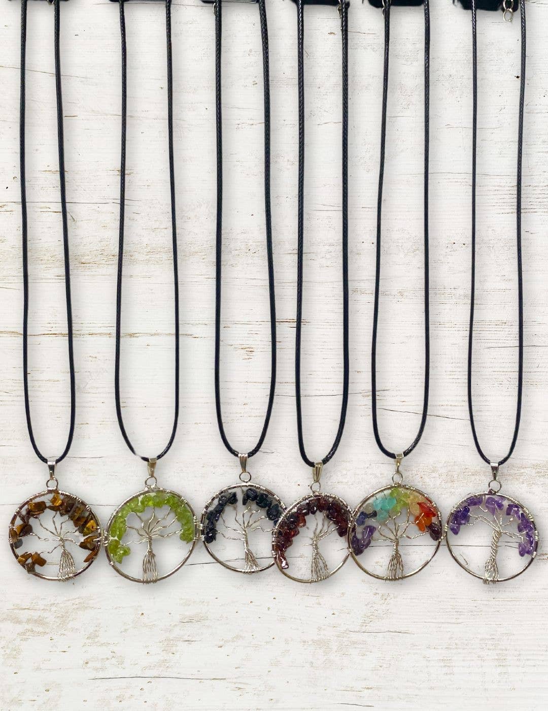 Six Silver Plated Tree of Life Stone Pendants, each featuring vibrant gemstone chips in different colors, are arranged in a row on a white wooden background. Each semi-precious pendant displays unique charm and beauty.