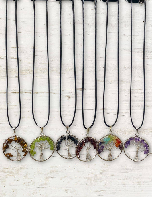 Six Silver Plated Tree of Life Stone Pendants, each featuring vibrant gemstone chips in different colors, are arranged in a row on a white wooden background. Each semi-precious pendant displays unique charm and beauty.