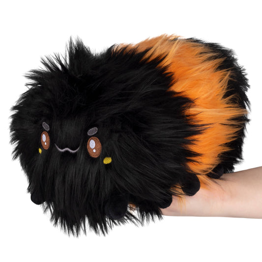 A hand holds the Mini Squishable Woolly Caterpillar, a plush toy with long black and orange fur, round eyes, and tiny legs—resembling the woolly caterpillars famed for predicting winter weather.