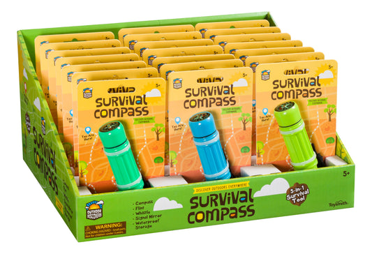 A display box holds multiple Outdoor Discovery Survival Compass packs in green or blue, each featuring a compass, flint, whistle, signal mirror, and waterproof storage. Suitable for ages 5 and up.
