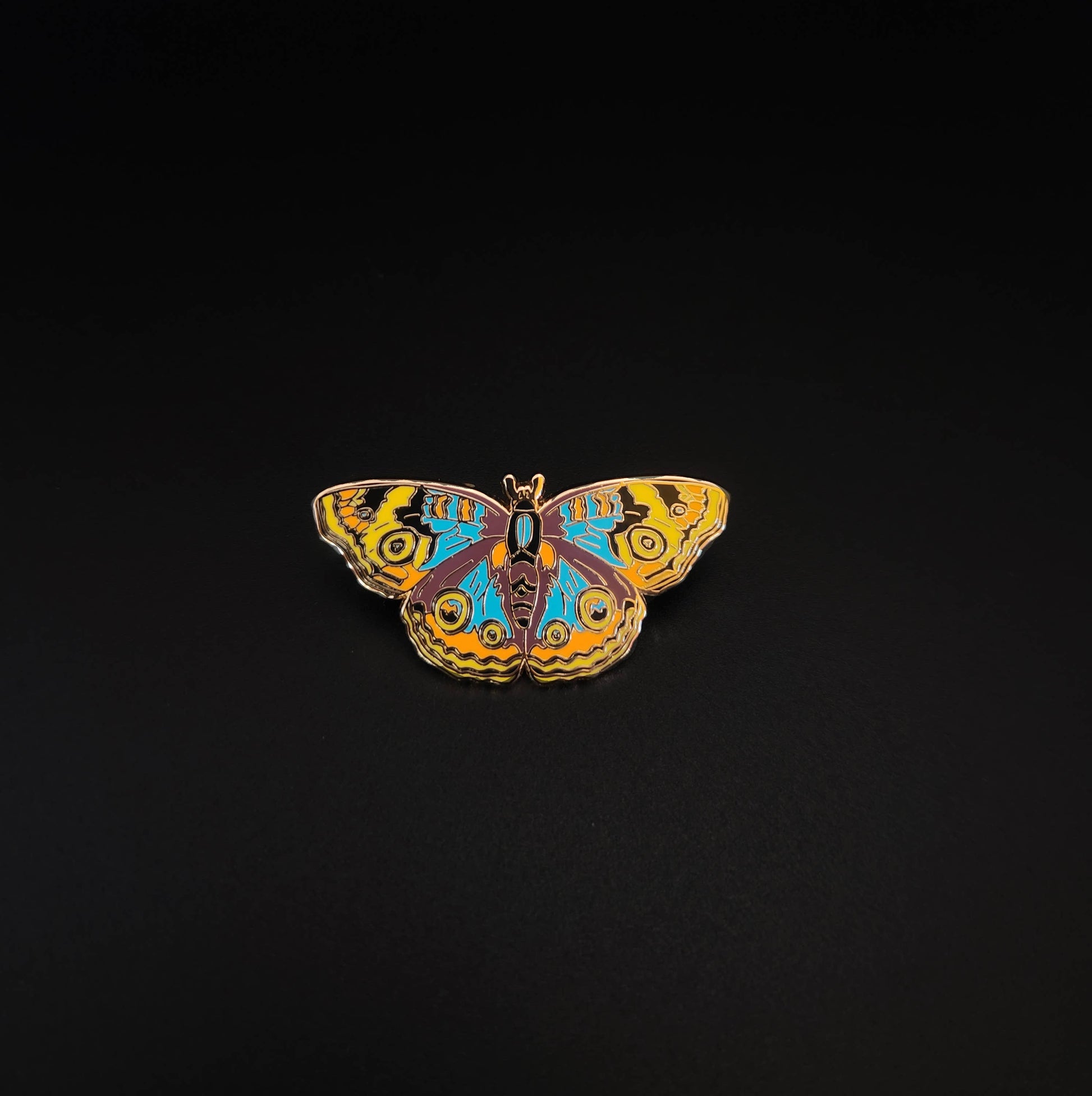 The Buckeye Butterfly Educational Insect Enamel Pin features vibrant yellow, blue, orange, and black patterns on a butterfly design set against a plain black background—making it an ideal gift for butterfly enthusiasts or a distinctive addition to any educational butterfly pin collection.