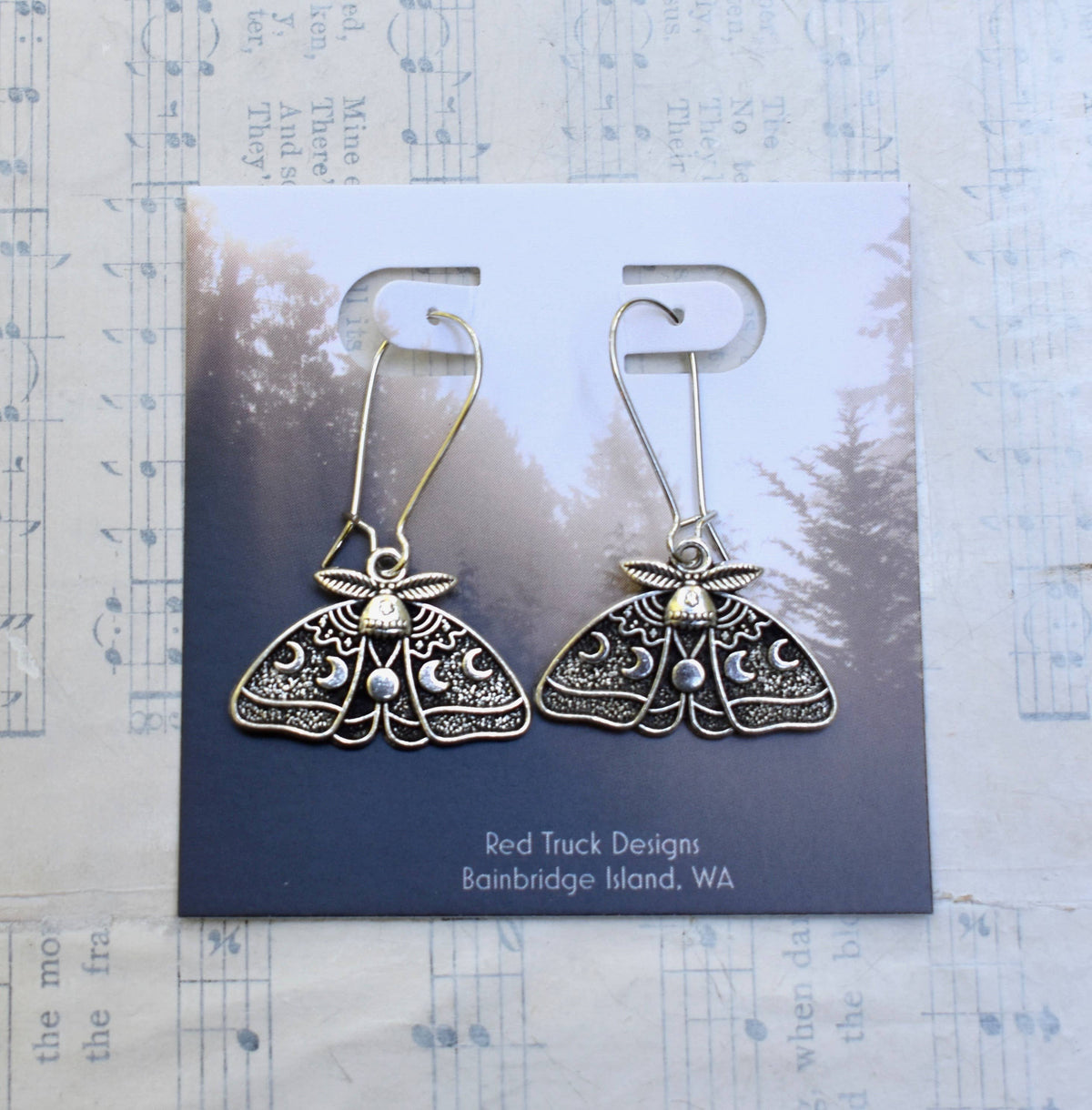 Luna Moth Earrings with intricate silver designs are displayed on a card featuring a forest background. Inspired by moonlit nights, these celestial earrings add an enchanting touch to any style. The display card is labeled, “Red Truck Designs, Bainbridge Island, WA,” and rests atop sheet music paper.