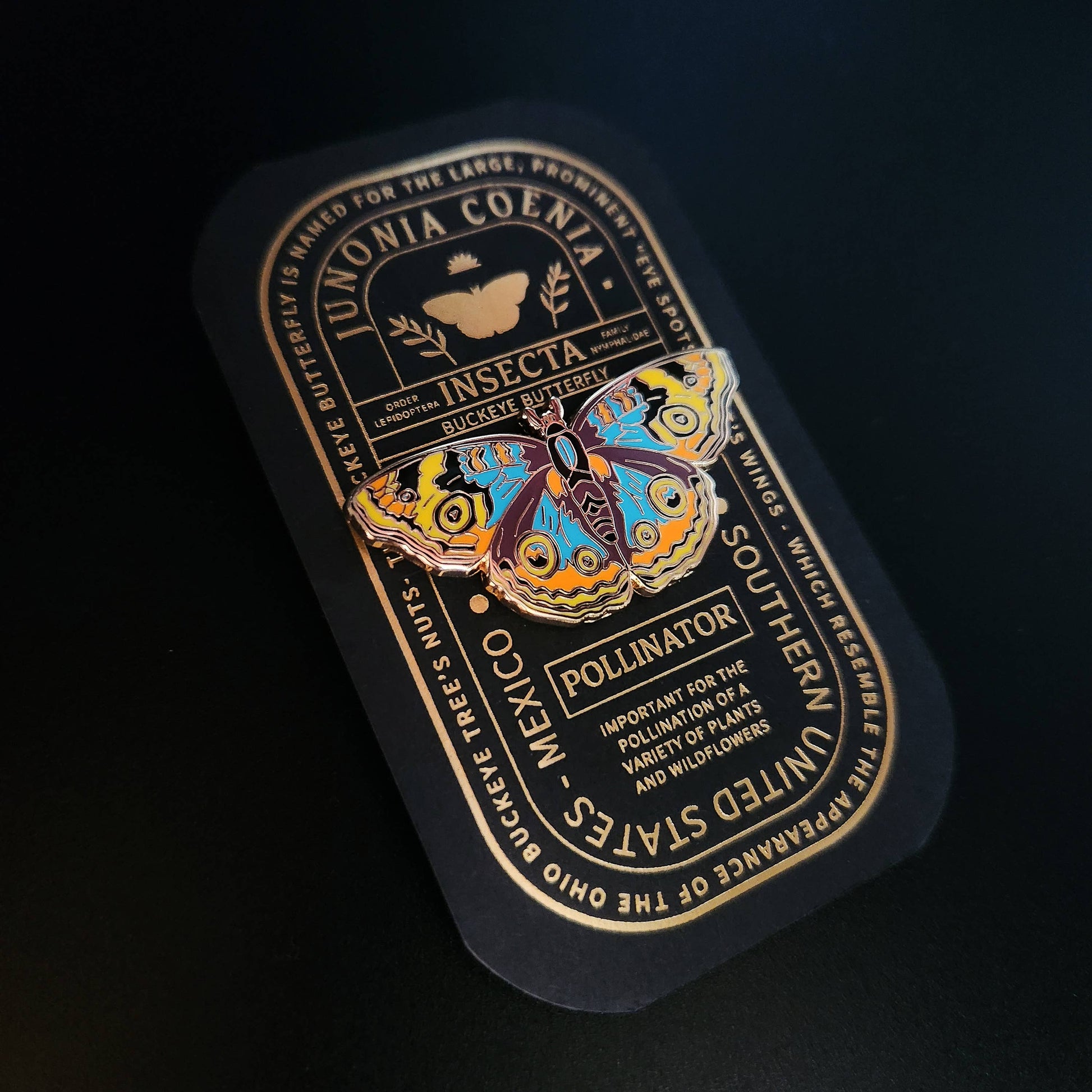 A vibrant butterfly illustration adorns a black card with gold text that shares interesting facts about the Buckeye Butterfly, such as its scientific name, habitat, and importance as a pollinator. The Buckeye Butterfly Educational Insect Enamel Pin makes an excellent gift for butterfly lovers or an educational accessory for nature enthusiasts.