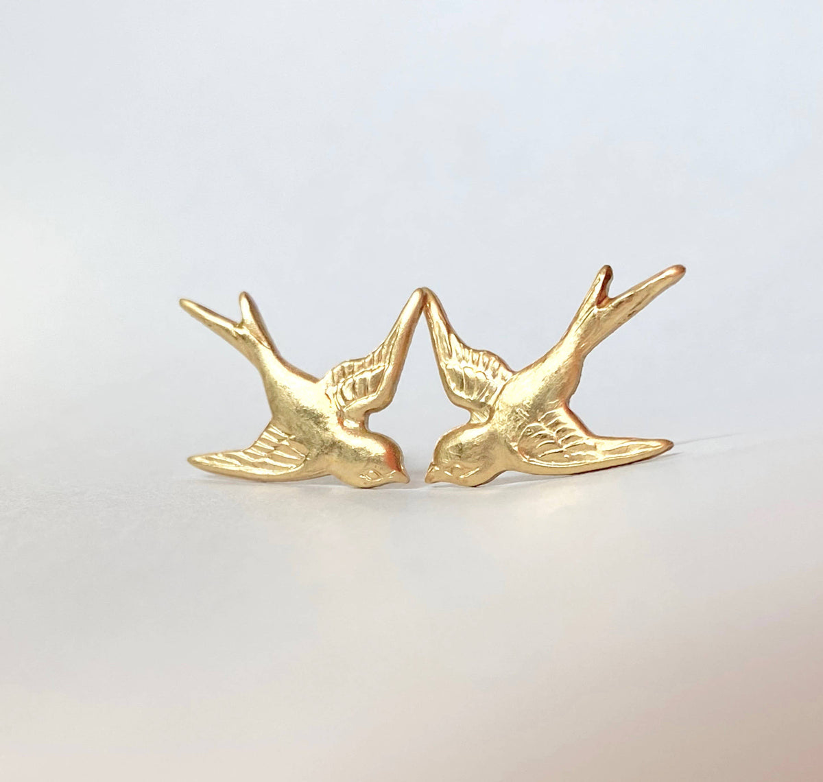 A pair of *Flying Sparrow Earrings* in golden brass, beautifully nature-inspired, face each other on a plain, light background—perfect woodland wedding earrings for those who adore delicate details.