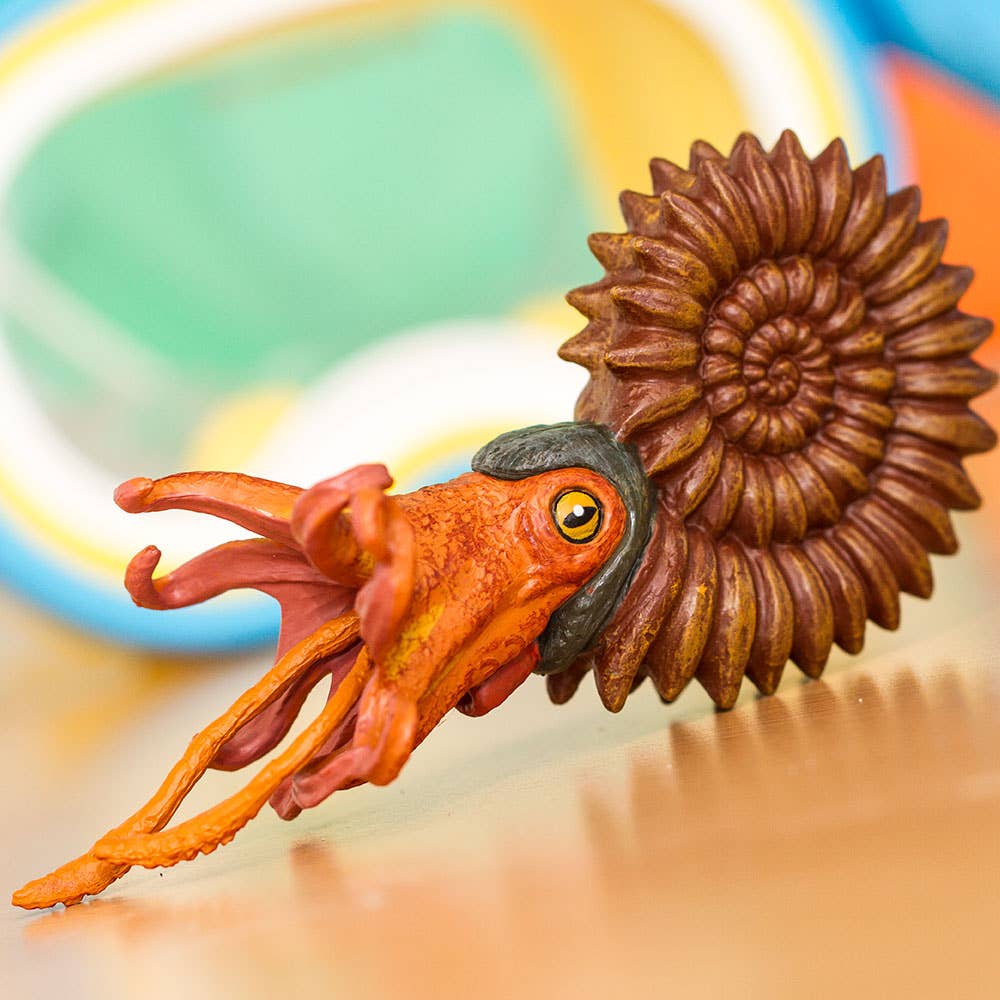 An Ammonite Figurine, intricately designed to resemble a real orange ammonite with spiral shell and tentacles, is displayed on a surface with blurred colorful objects in the background.