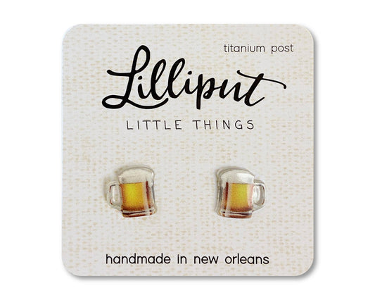 Introducing the Beer Mug Earrings: a pair of small novelty earrings handcrafted in New Orleans and shaped like miniature beer mugs filled with amber liquid. Displayed on a card labeled Lilliput Little Things, these earrings feature hypoallergenic titanium posts—making them perfect for sensitive ears.