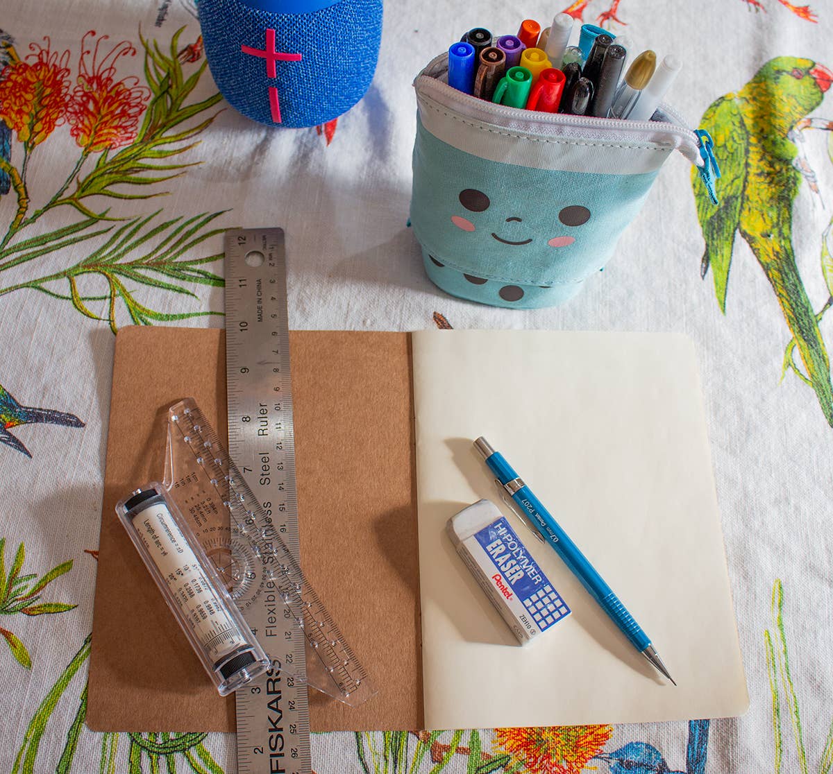An open Sketch Book Egyptian Horus & Maat - 6" x 8" with a blue mechanical pencil, eraser, and clear ruler rests on a floral tablecloth with birds. Nearby: metal ruler, battery, blue marker case, and blue speaker in Cairo artisan style.