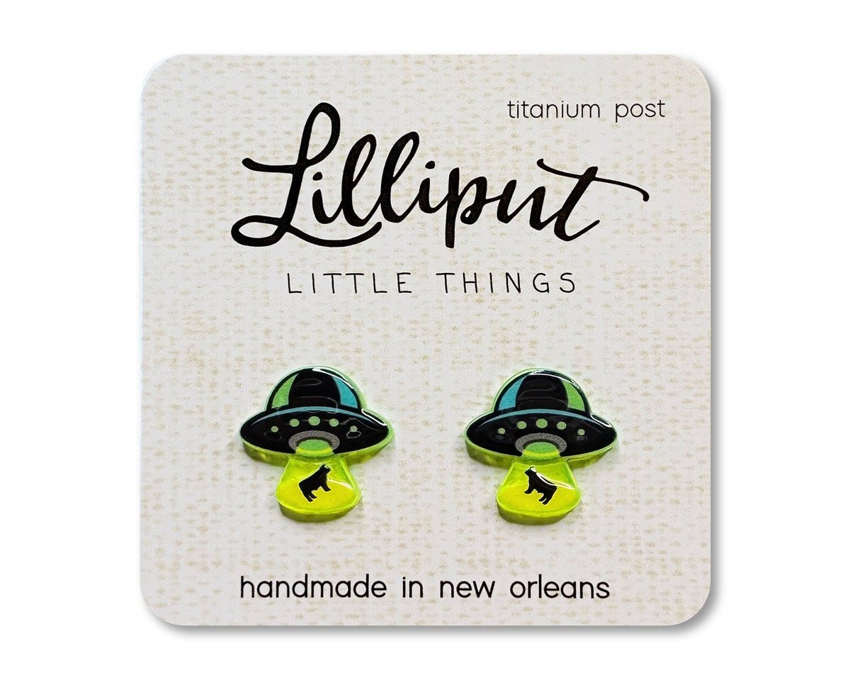 A pair of UFO Earrings crafted from titanium and shaped like flying saucers beaming up a black cat, presented on a Lilliput Little Things card marked hypoallergenic and handmade in New Orleans.