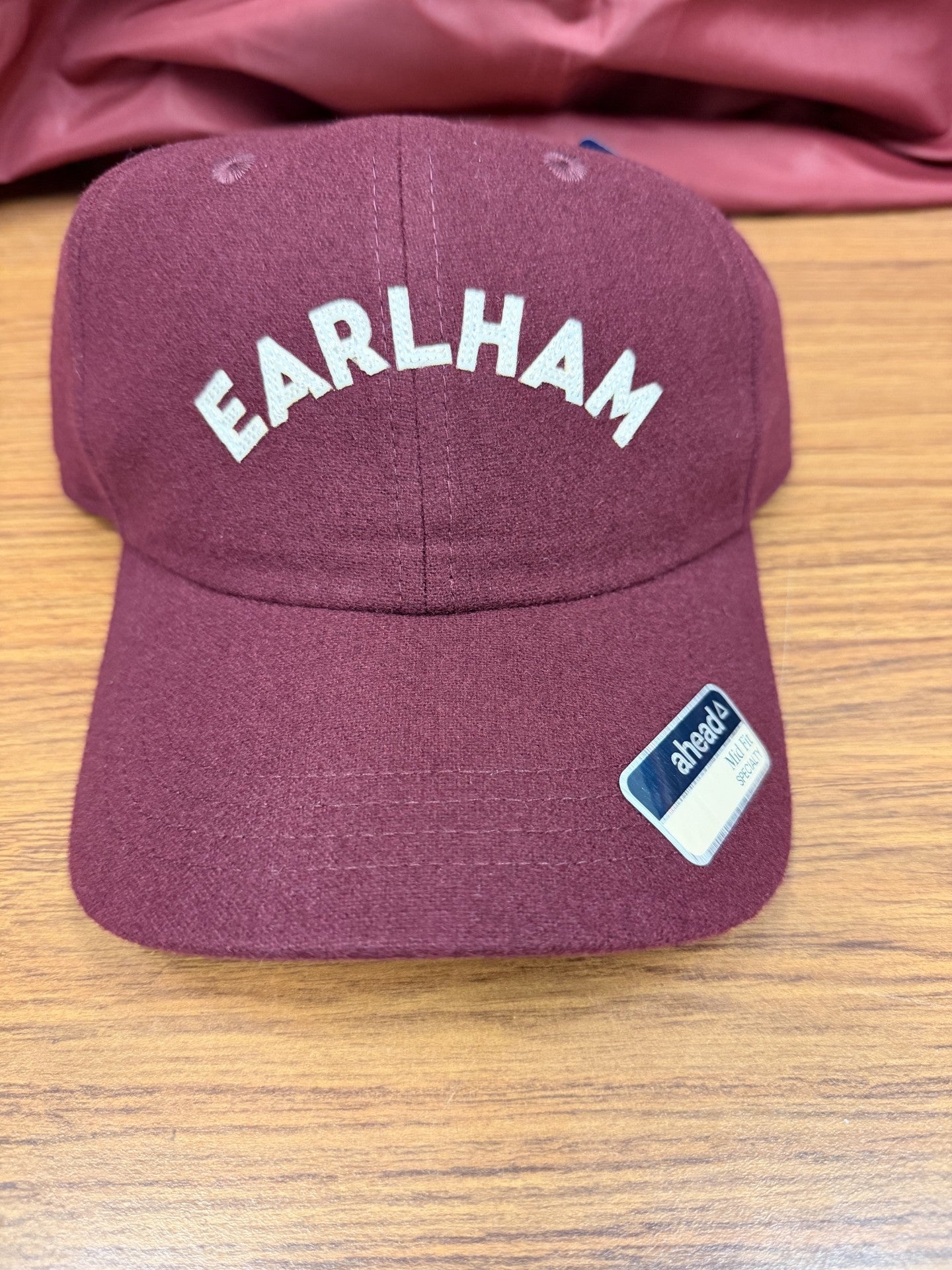A Clubhouse Lightweight Wool Hat in maroon, featuring "EARLHAM" embroidered in white on the front, rests on a wooden surface with a small "ahead" sticker displayed on the brim.