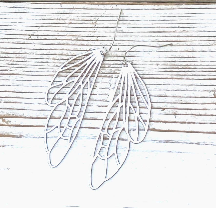 A pair of Silver Lace Cicada Wings earrings, shaped like delicate insect wings, rests on a light wooden surface with horizontal lines.