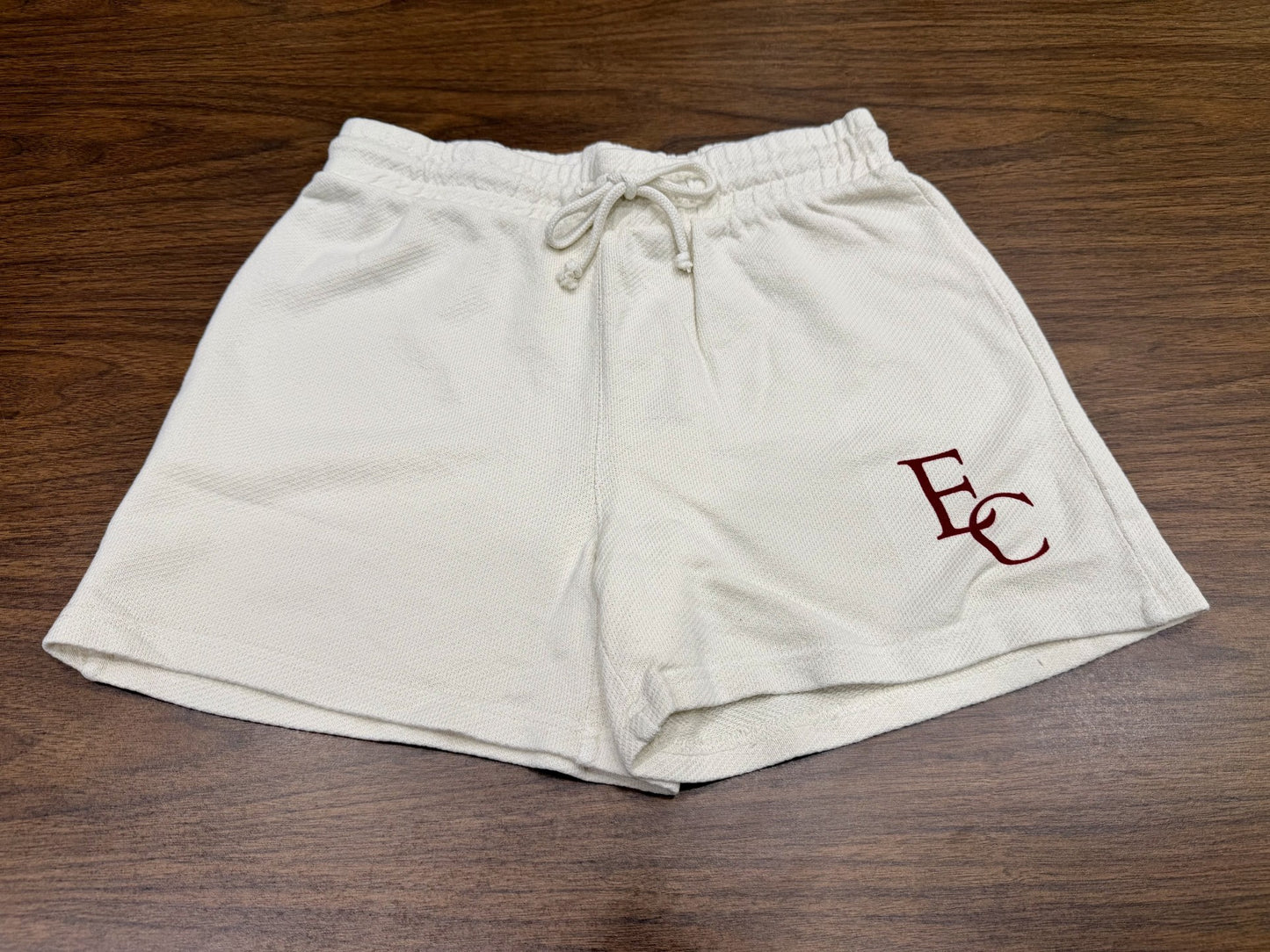 French Terry Shorts with a drawstring waistband, in white, displayed on a wooden surface. Dark red EC initials are printed near the hem of the left leg.
