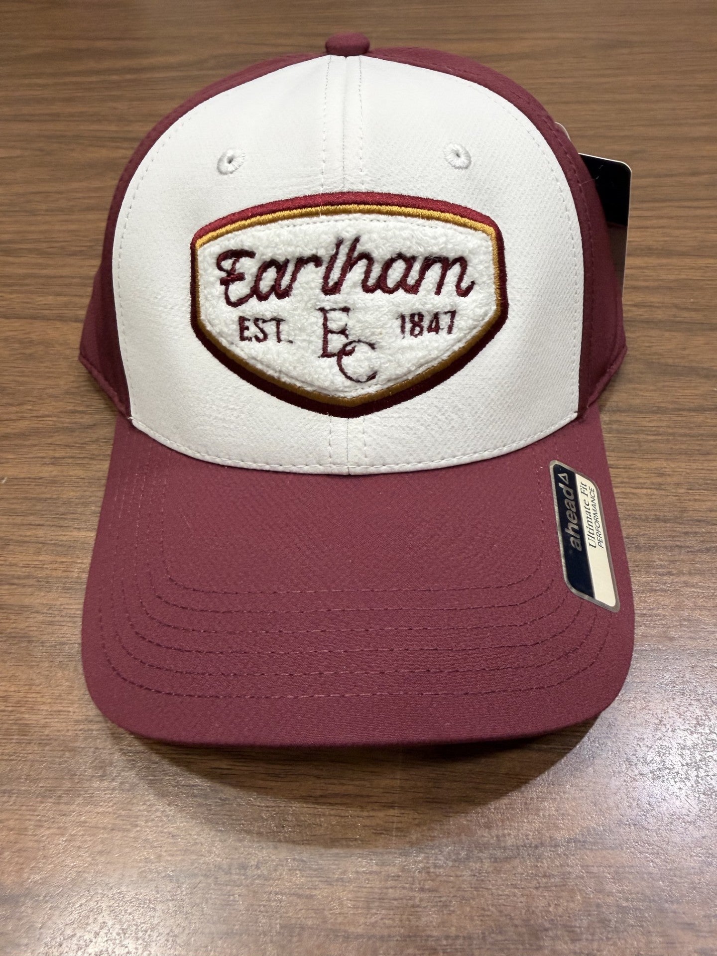 An AeroSphere Lightweight Velcro Hat in maroon and white, featuring an embroidered patch reading "Earlham EC EST 1847," rests on a wooden surface. A sticker on the brim displays the AHEAD brand.