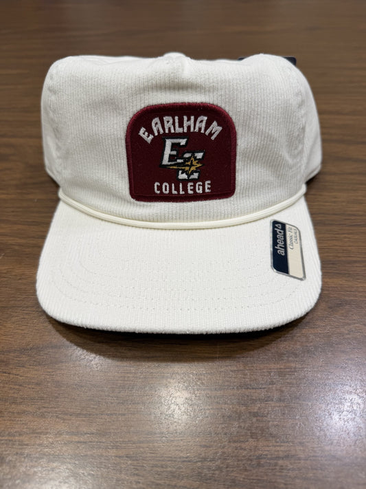 A Corduroy Snapback Rope Hat featuring a maroon and white Earlham College patch and a small black and white arcaform tag rests on a wooden surface.