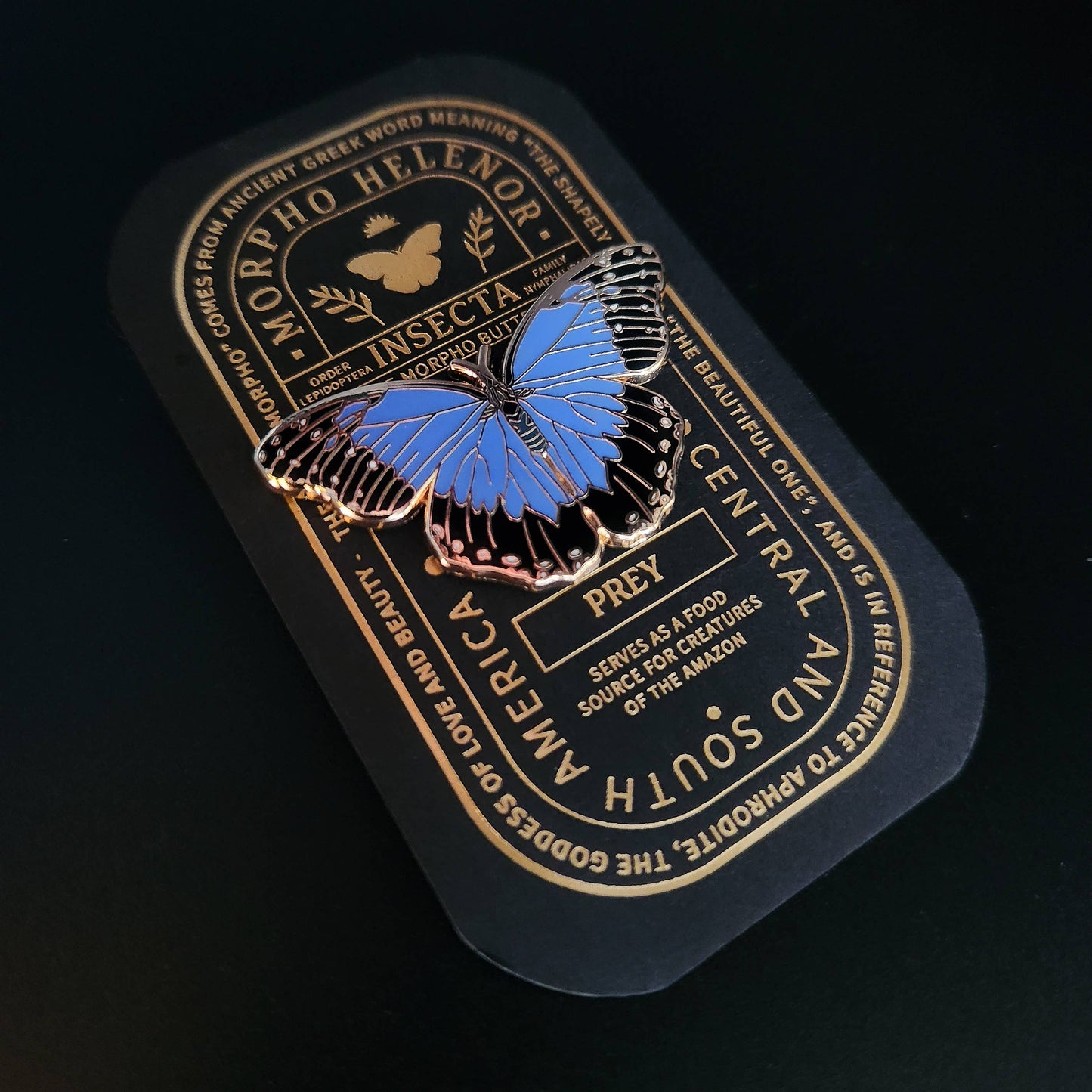 The Blue Morpho Butterfly Educational Insect Enamel Pin is displayed on a decorative black and gold card featuring information about the Morpho Helenor butterfly, including its insect classification and role as prey in South America—making it a beautiful nature-themed gift for any butterfly enthusiast.
