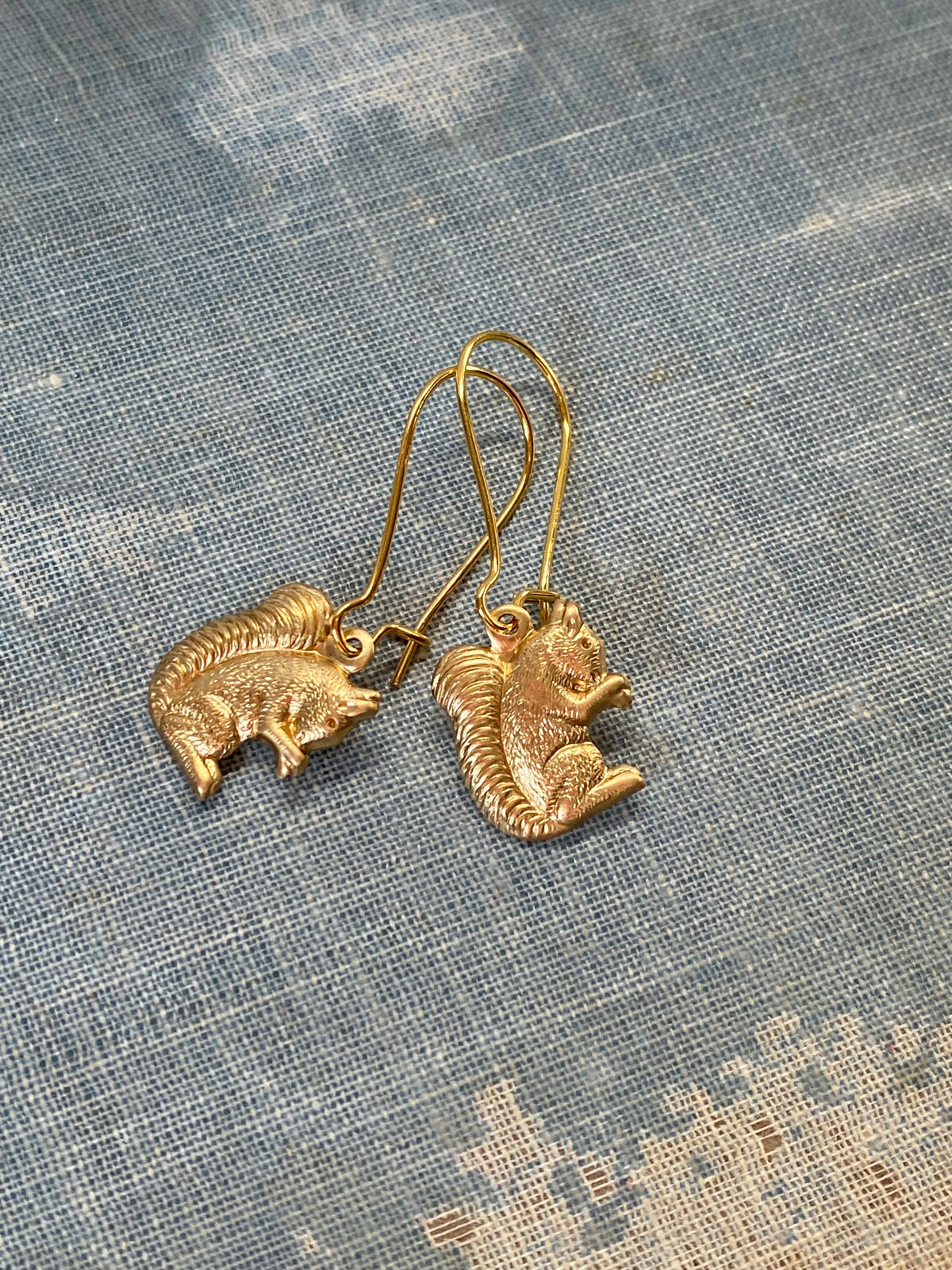 A pair of Woodland Forest Squirrel Earrings, delicately shaped like charming forest creatures, rests on a blue and white textured fabric surface.