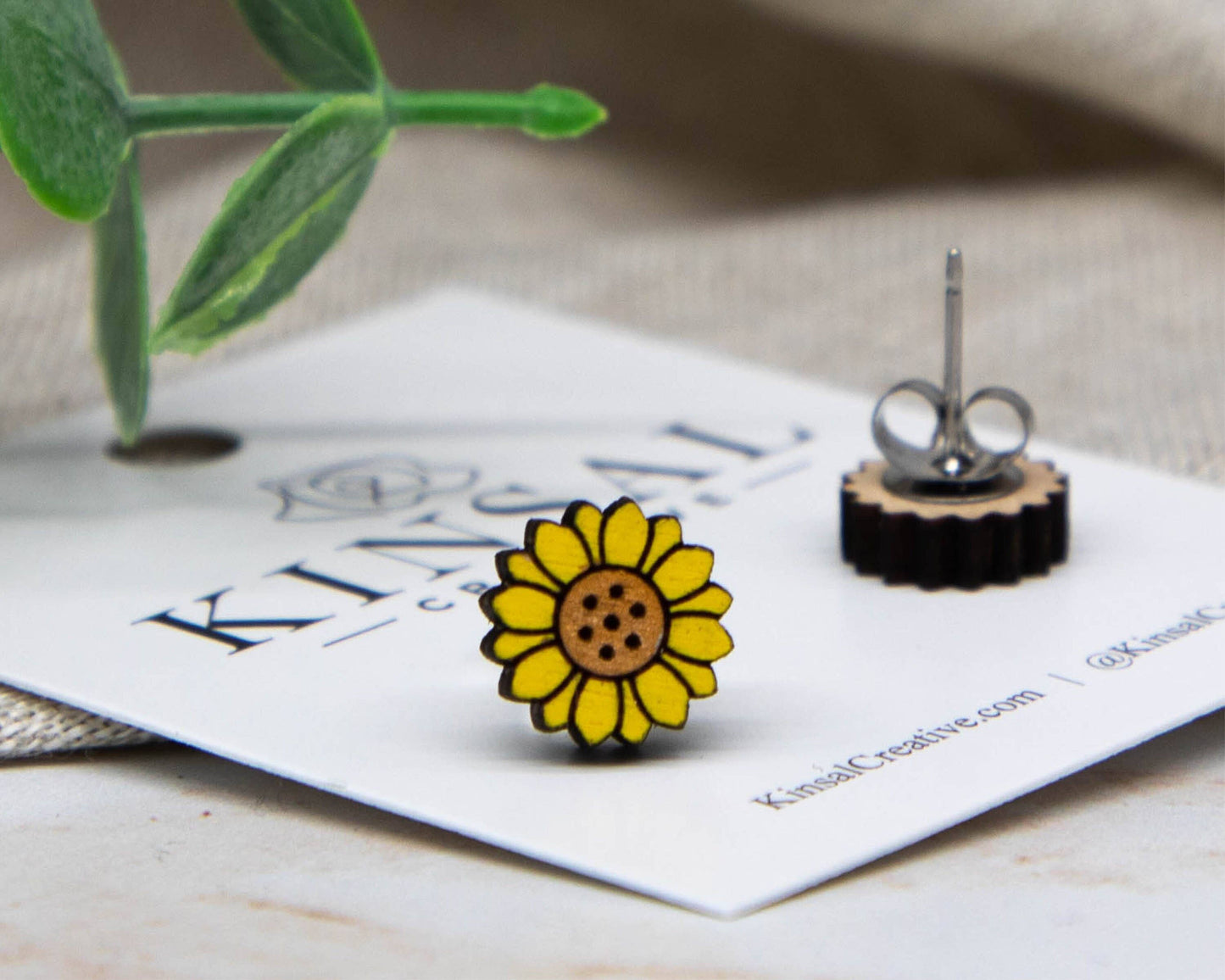 A close-up of Sunflower Wood Stud Earrings, shaped like yellow sunflowers, is shown on a white card with Kinsal branding. One earring lies flat beside it. Green leaves and neutral fabric accent this handmade jewelry.