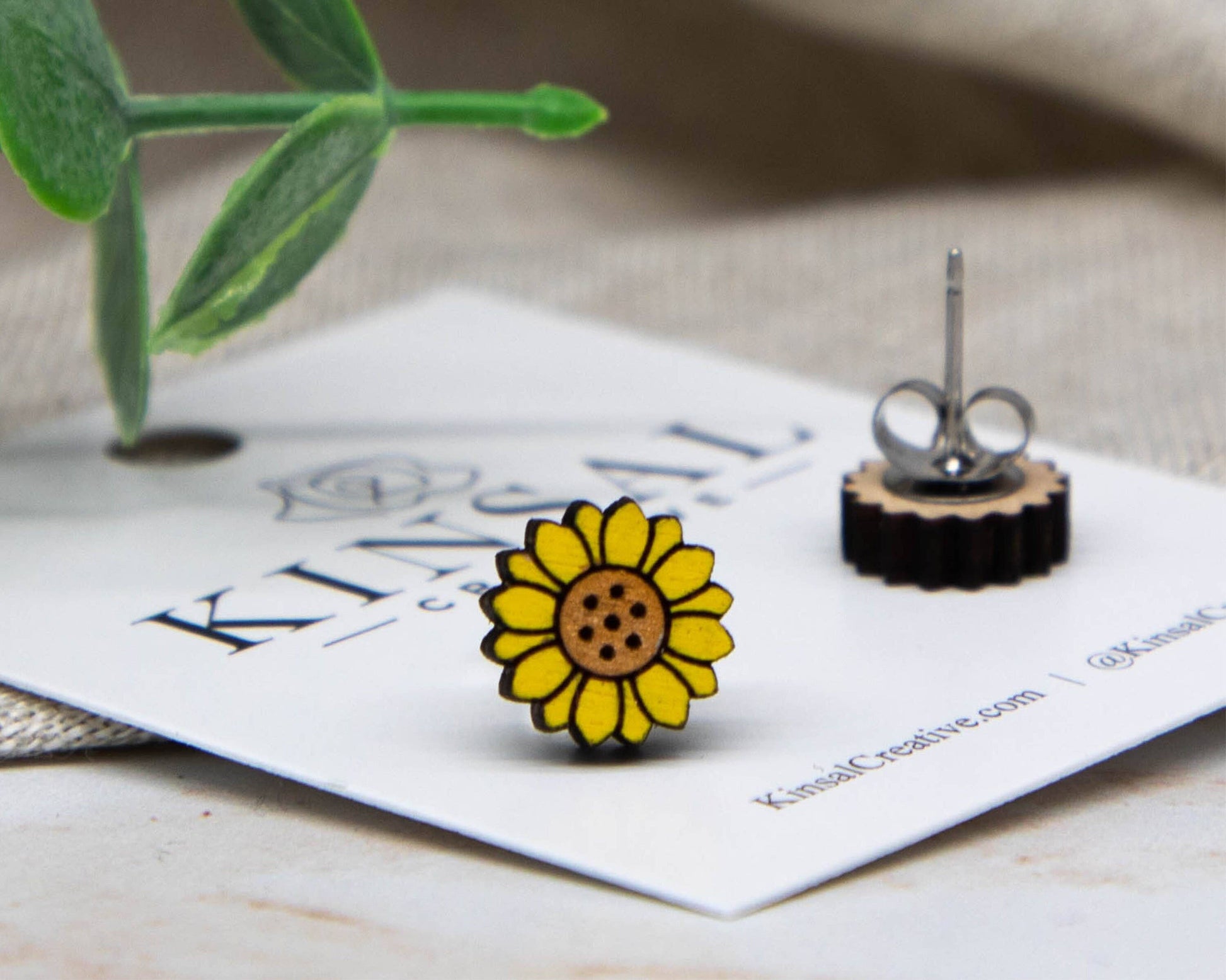 A close-up of Sunflower Wood Stud Earrings, shaped like yellow sunflowers, is shown on a white card with Kinsal branding. One earring lies flat beside it. Green leaves and neutral fabric accent this handmade jewelry.