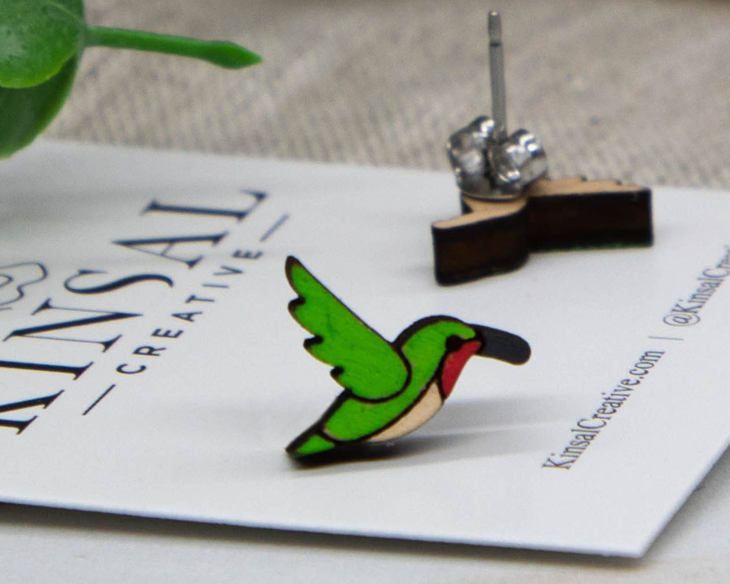 The Hummingbird Wood Stud Earrings feature a small, colorful design with green wings and a red accent, displayed on a branded card. Matching earring and stainless steel back are shown in the background—ideal for handmade jewelry lovers.