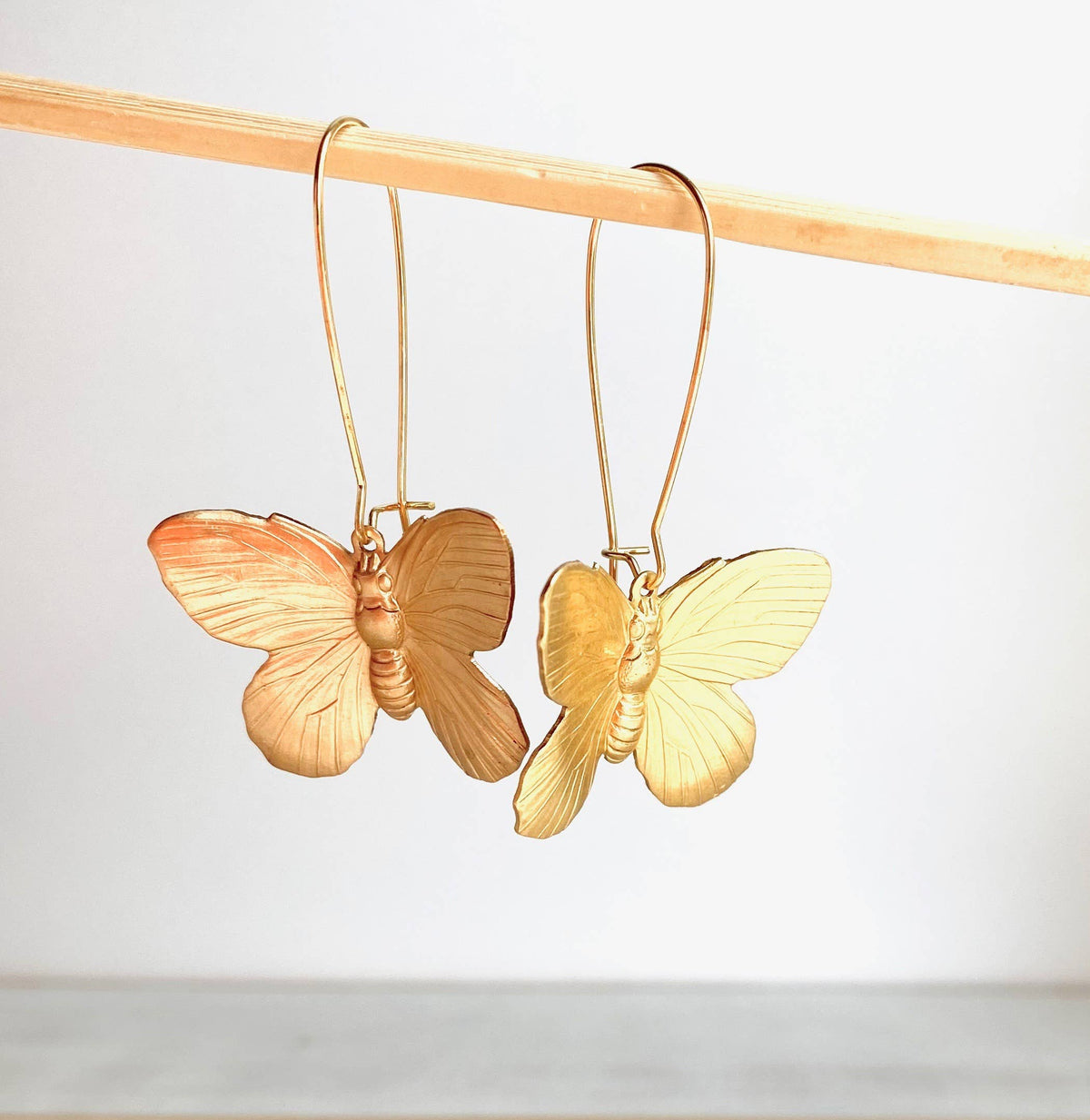 Butterfly Earrings hang from a thin wooden rod, displayed against a plain white background. These statement earrings showcase delicate, detailed wings and elegant curved hooks, beautifully crafted from golden brass for a touch of timeless elegance.