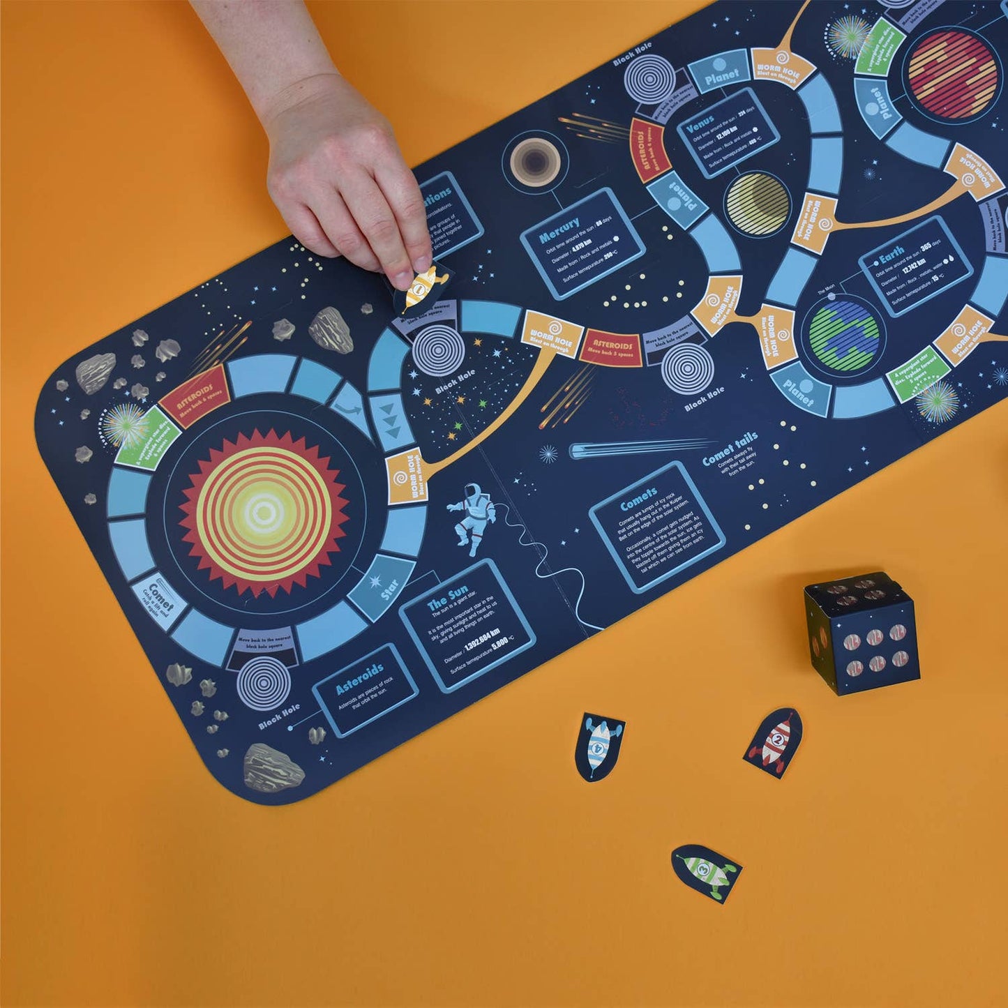A hand moves a piece on the "Create Your Own Solar System" game—featuring colorful paths, planets, and cards on an orange surface. Other pieces and a black die are nearby, enhancing this space-themed board game adventure.